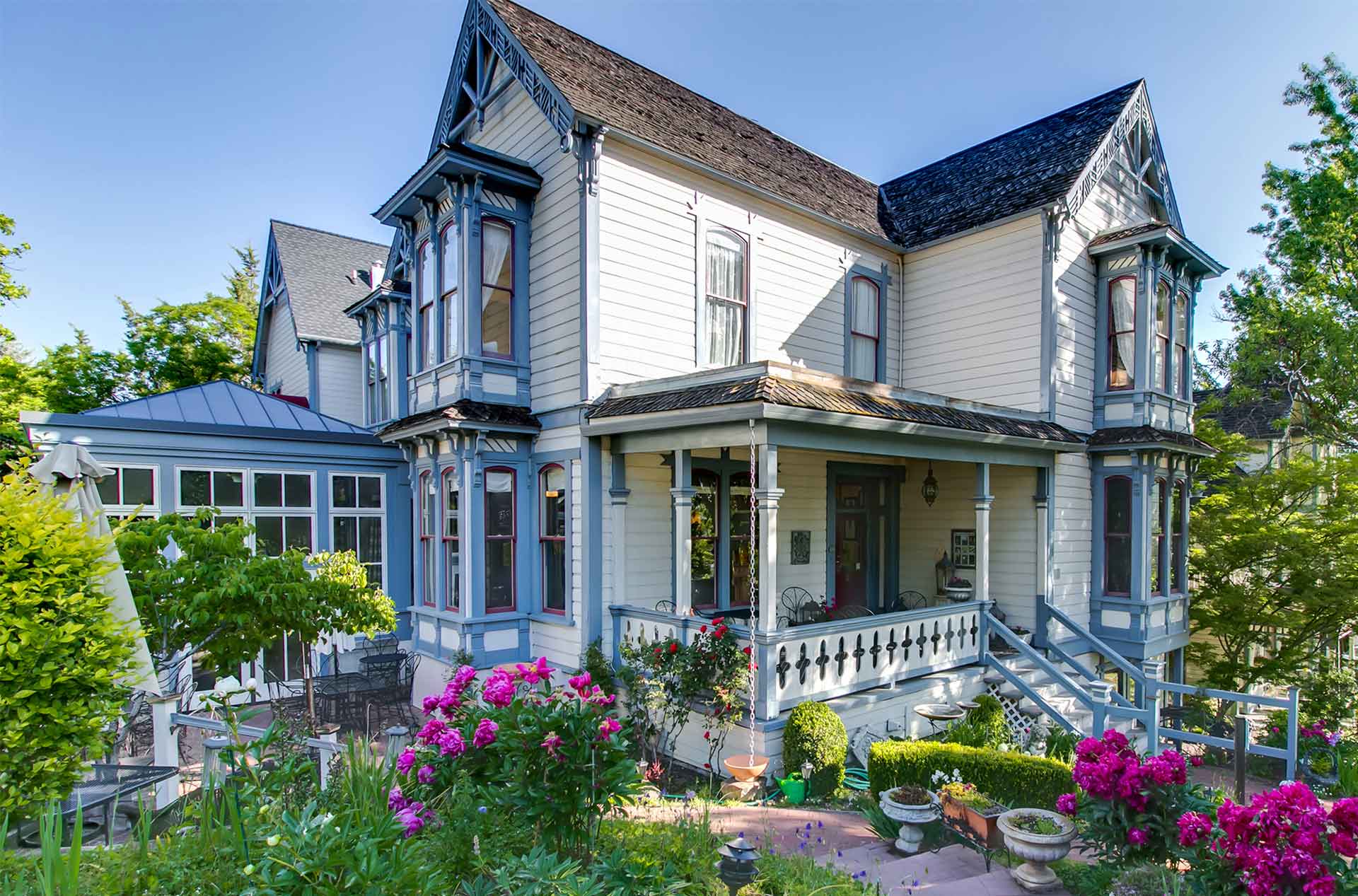 Victorian-style inn with ornate blue and white trim, a covered porch, and lush gardens. Bay windows and gabled roofs add to its charm under a clear blue sky.