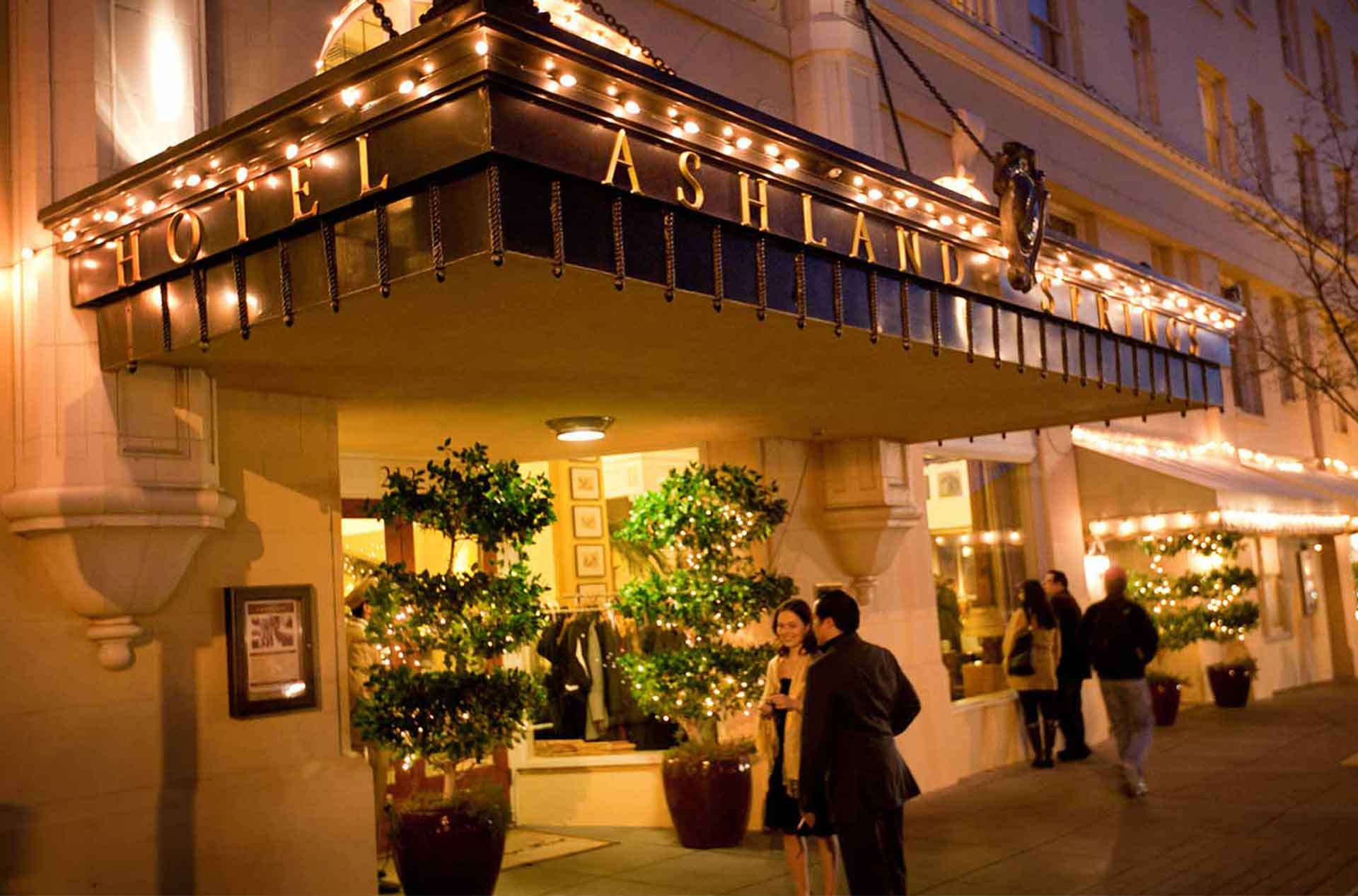The illuminated entrance of Ashland Springs Hotel at night, featuring warm string lights, potted trees, and guests in formal attire chatting near the front door.