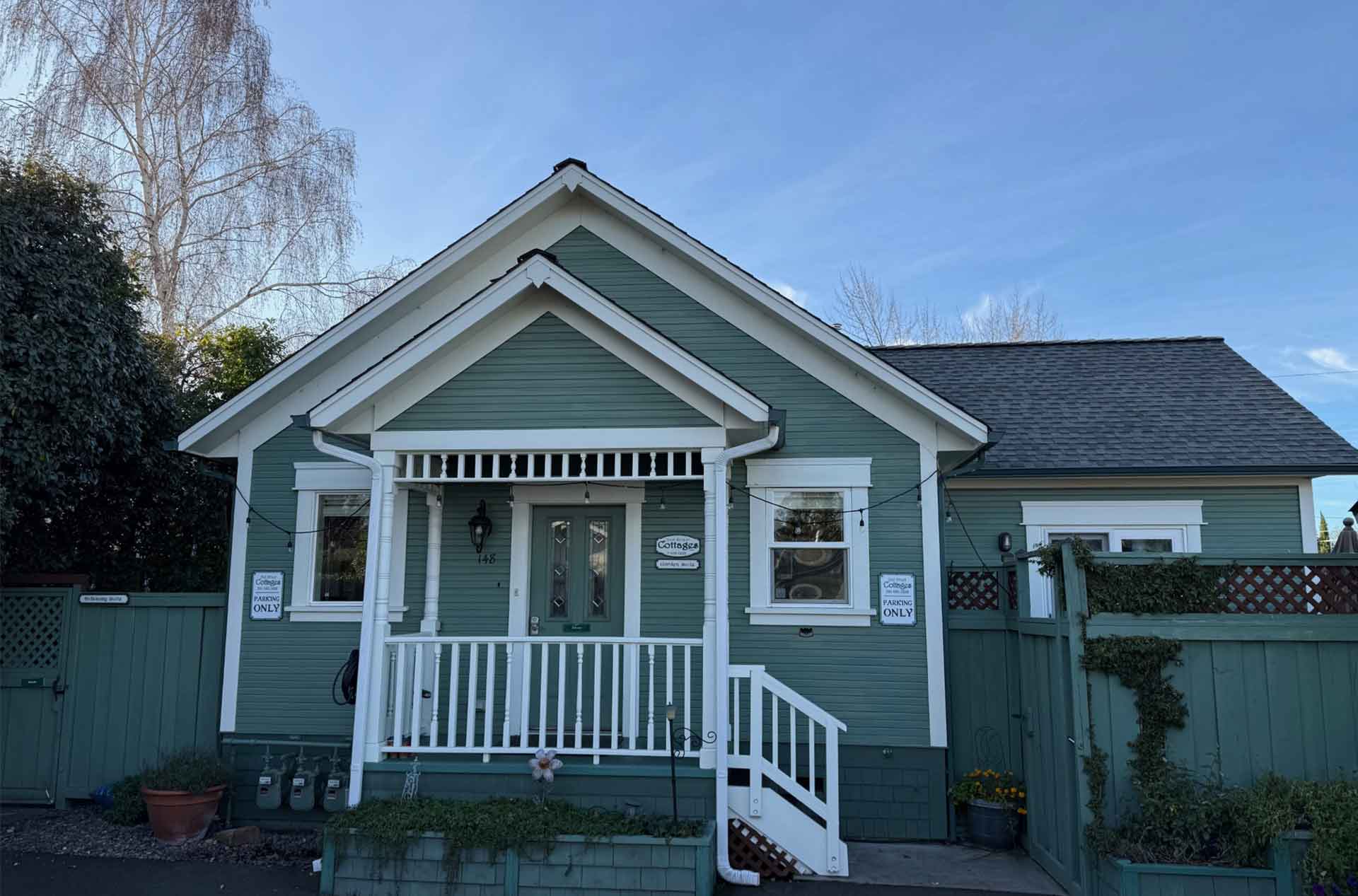 A small green cottage with white trim, a front porch, and a fenced yard under a clear blue sky.