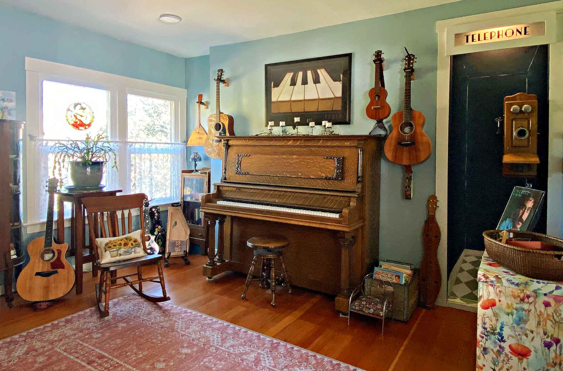 A cozy room featuring a vintage piano, several guitars and string instruments hanging on the walls, a rocking chair, and a telephone booth. There's a window with sheer curtains, a potted plant, and colorful decor, including a patterned rug and a basket.