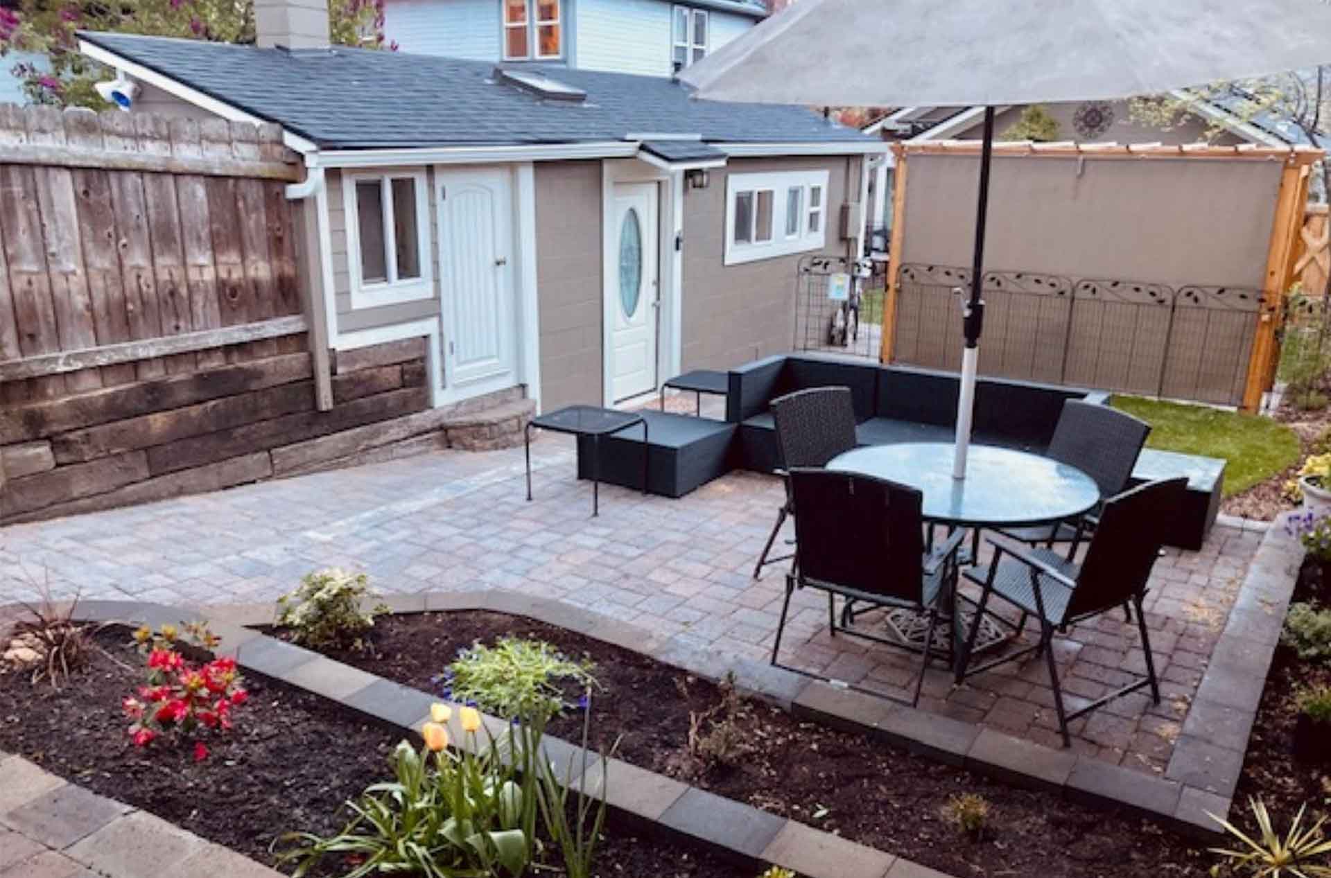A cozy backyard patio with a table, chairs, and a sectional couch, surrounded by flower beds and a wooden fence. A small shed is visible in the background.