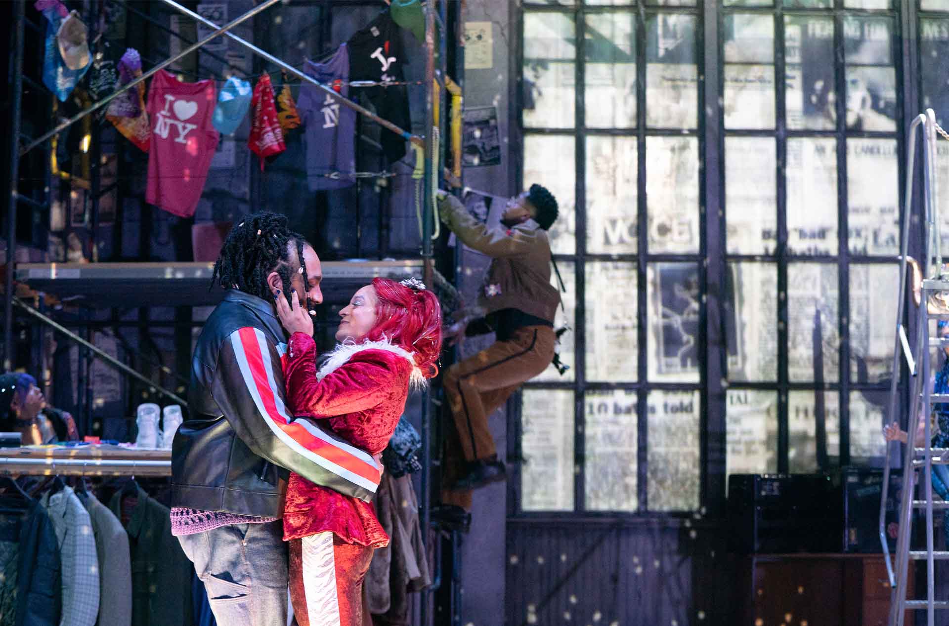 Two people embracing and looking at each other while someone climbs ladder in the background.
