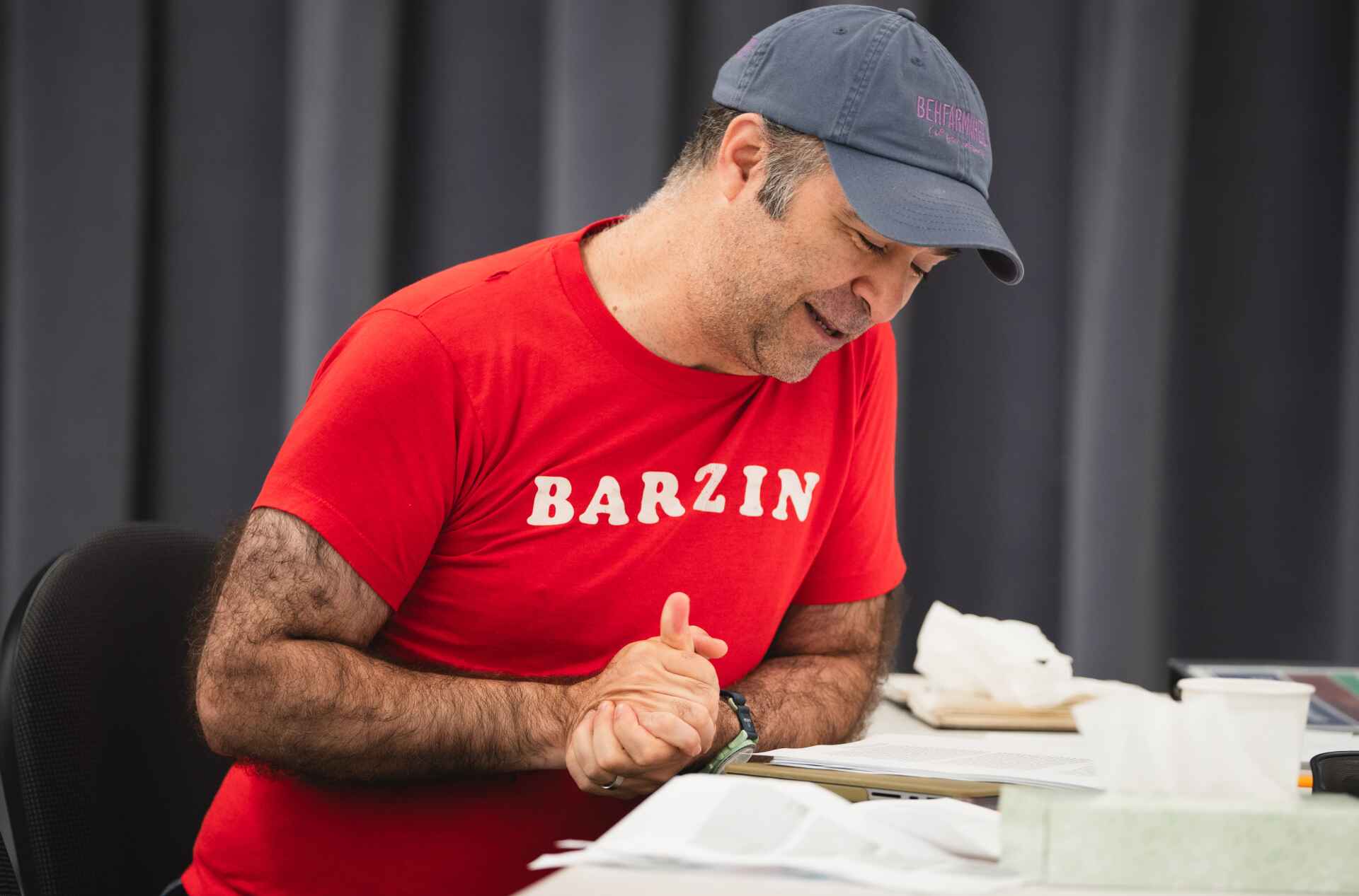 A person in a red BARZIN shirt and a blue cap is sitting at a table, reading a book with a tissue box and papers nearby.