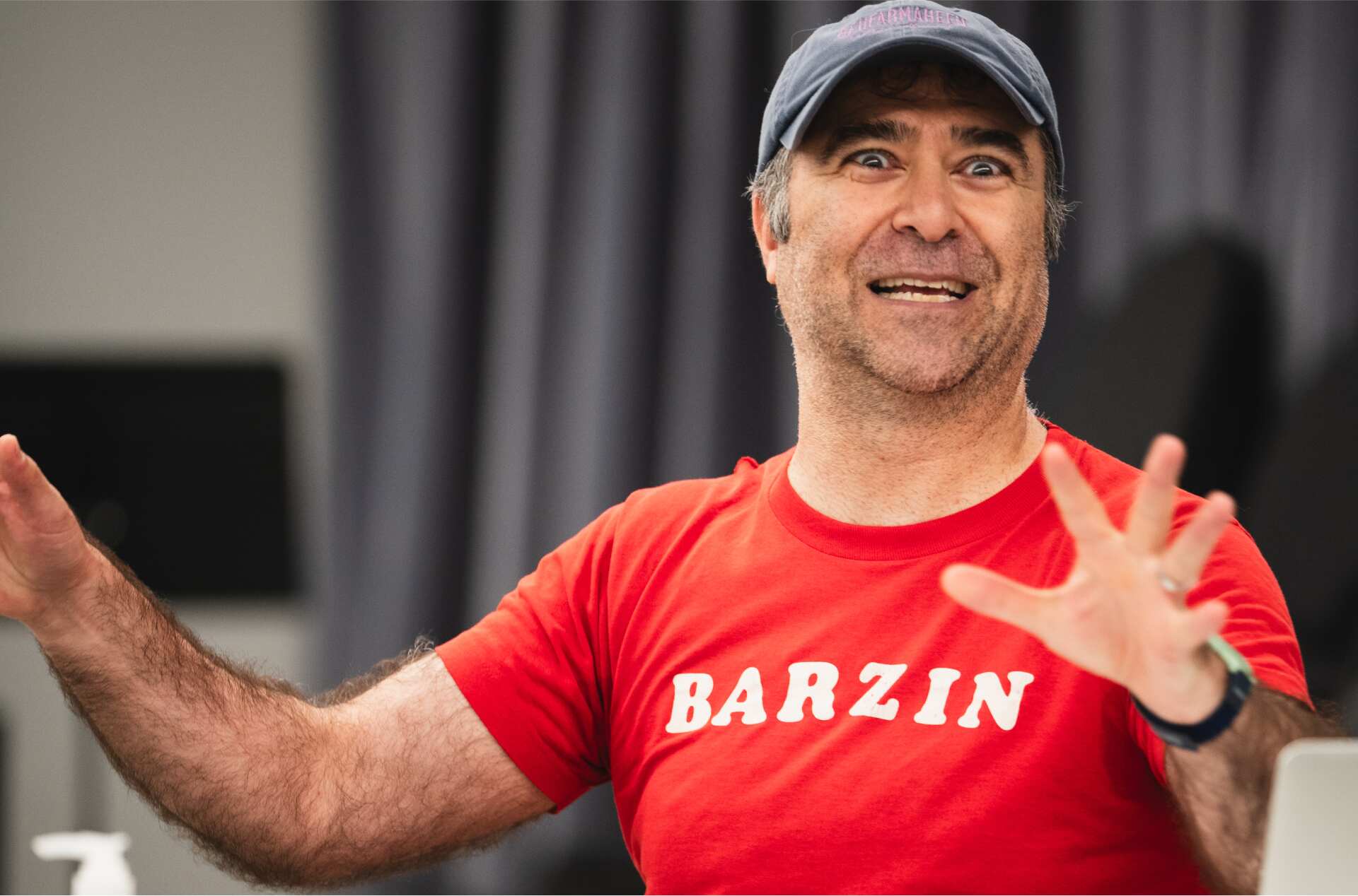 A person wearing a blue cap and a red T-shirt with "BARZIN" on it gestures animatedly with both hands. They are seated at a table with items such as a hand sanitizer bottle, a paper cup, and papers. A curtain is visible in the background.