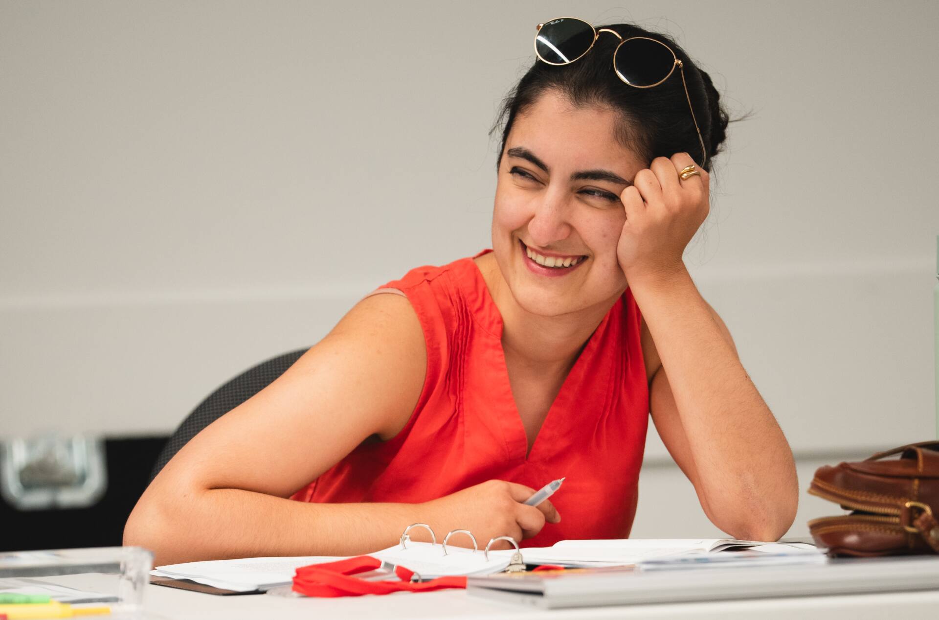 A person in a red sleeveless top sits at a table with papers, pens, and a pair of glasses. They are smiling, resting their head on their hand, with sunglasses perched on their head.