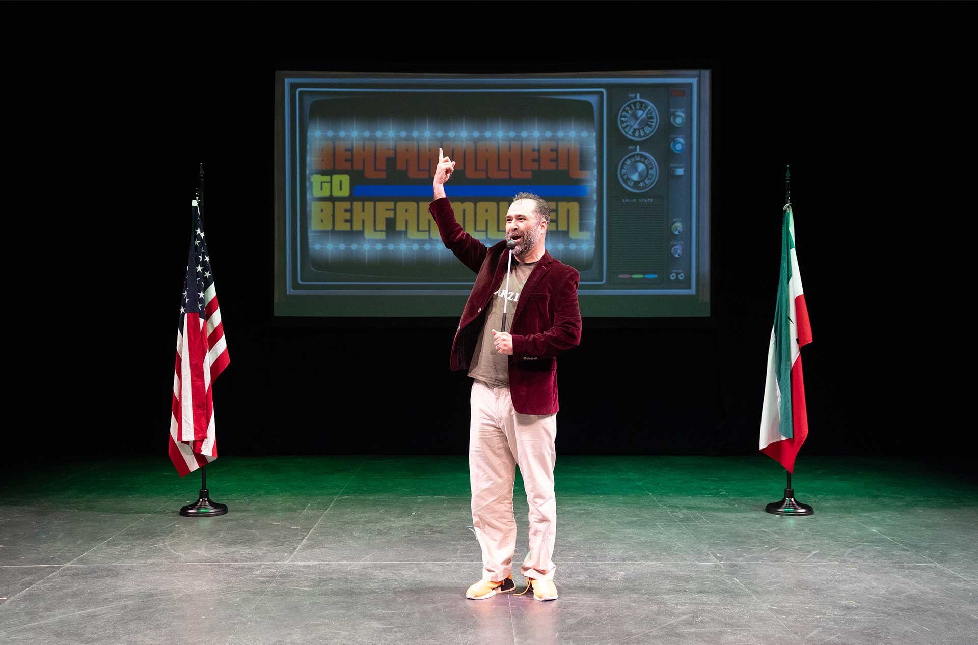 A person stands on stage between two flags, one American and one Iranian, gesturing upward with one finger. A screen behind displays the text "Behfarmeheen" in bold, colorful letters.