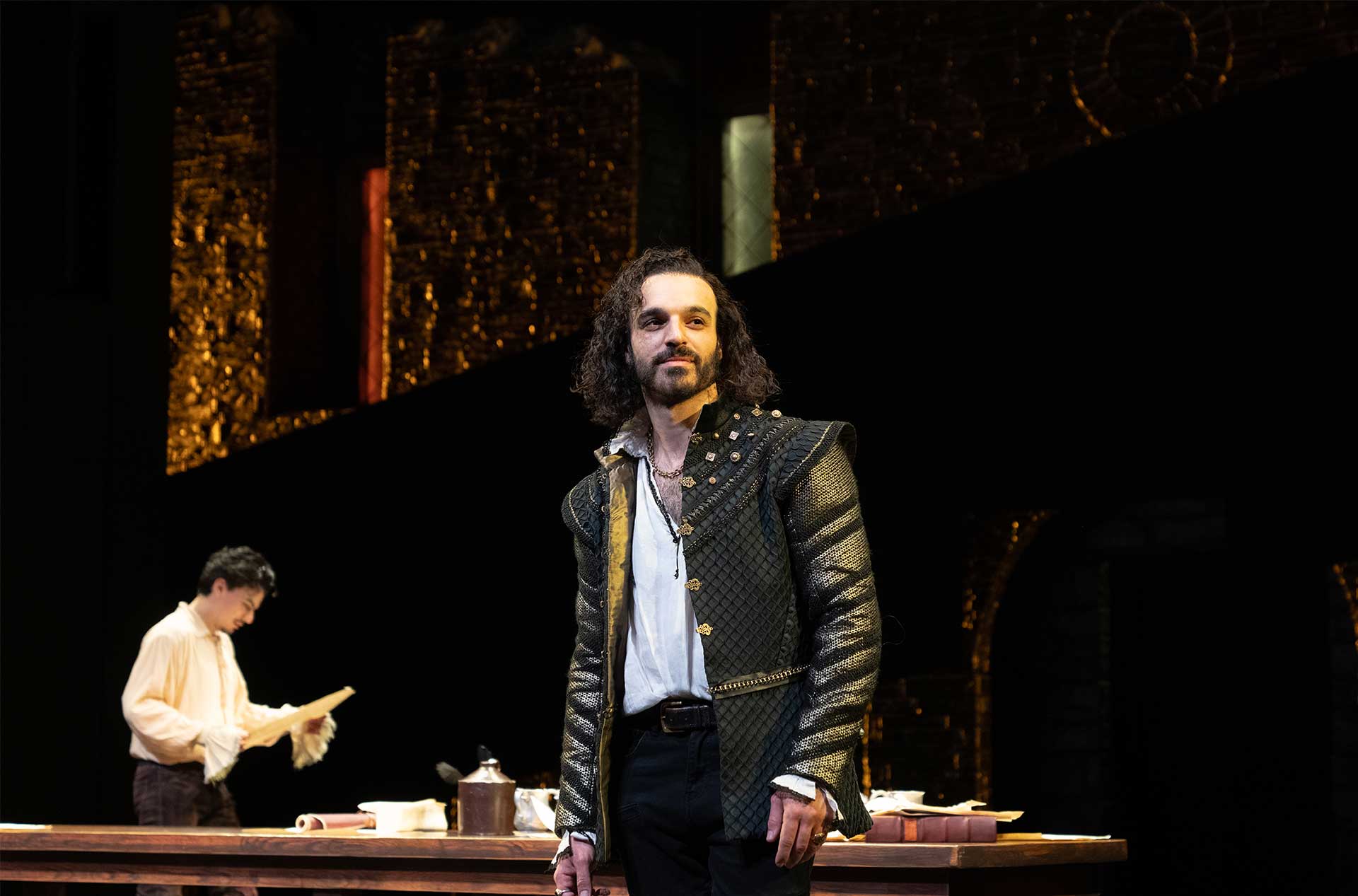 A person with long curly hair and a medieval-style outfit stands in the foreground, looking ahead confidently. In the background, another person is reading a document at a table set with various items, under warm, dramatic lighting.