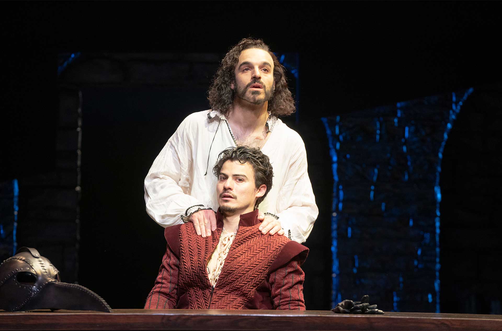 A person with long curly hair stands behind another seated person, resting their hands on the seated person's shoulders. The seated person has short dark hair and is wearing a red, quilted medieval-style outfit. Both are looking in the same direction with serious expressions.