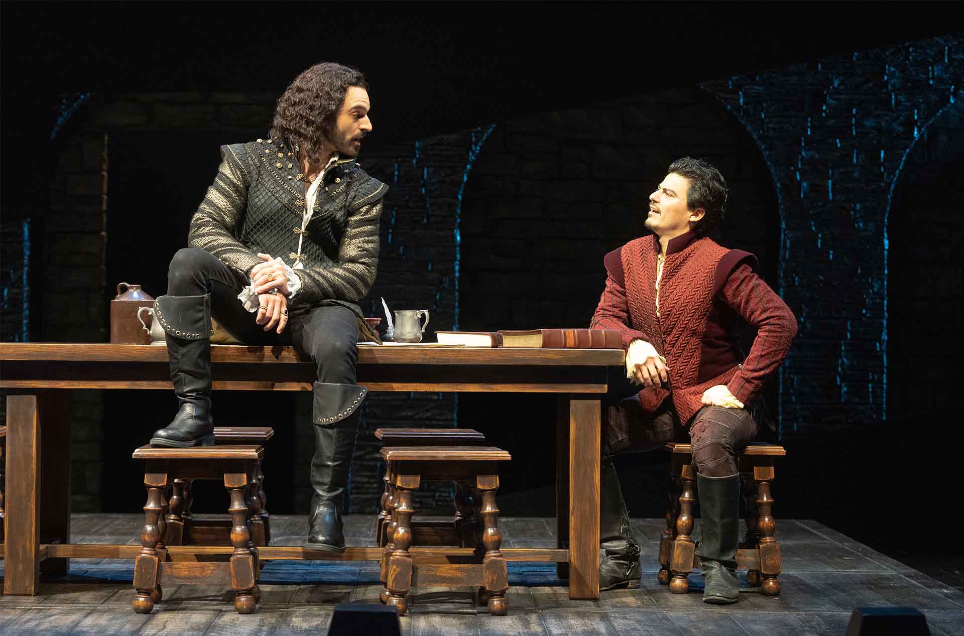 Two people in medieval-style outfits are at a wooden table. One person with long curly hair is sitting on the table, while the other person, with short dark hair, is seated on a stool and looking up at the first person. The table has books and metal cups on it.