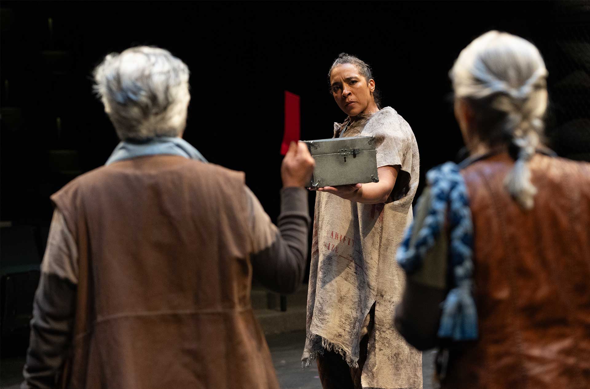 Clad in a ragged cloak, an individual holds out a small metal box towards another who is holding a red cloth. The scene suggests a significant exchange or revelation with intense expressions on both faces.