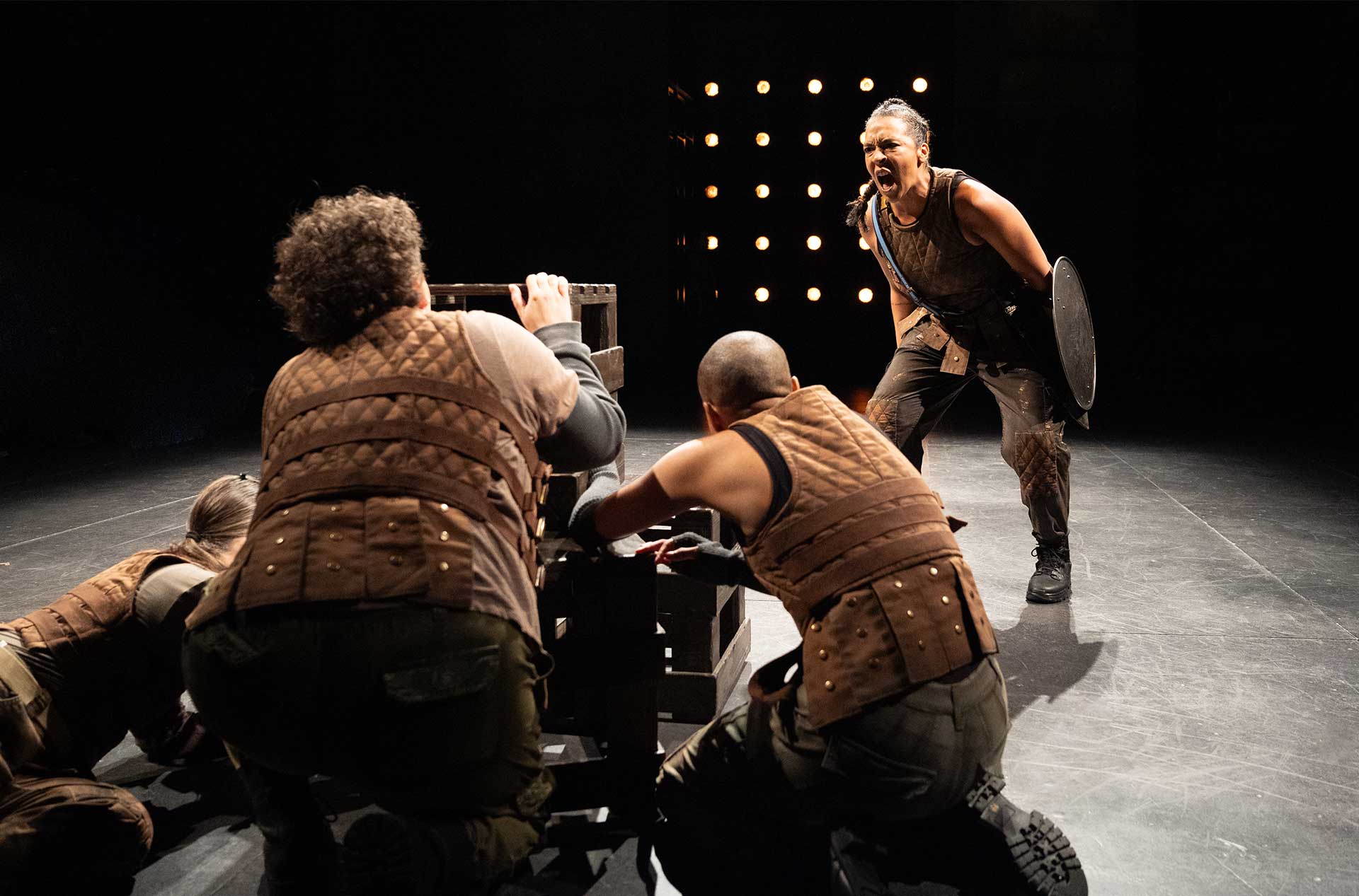 Three individuals in brown quilted vests crouch around a wooden structure on stage while another stands shouting passionately and holding a round shield. The scene is illuminated by a backdrop of stage lights creating a dramatic atmosphere.