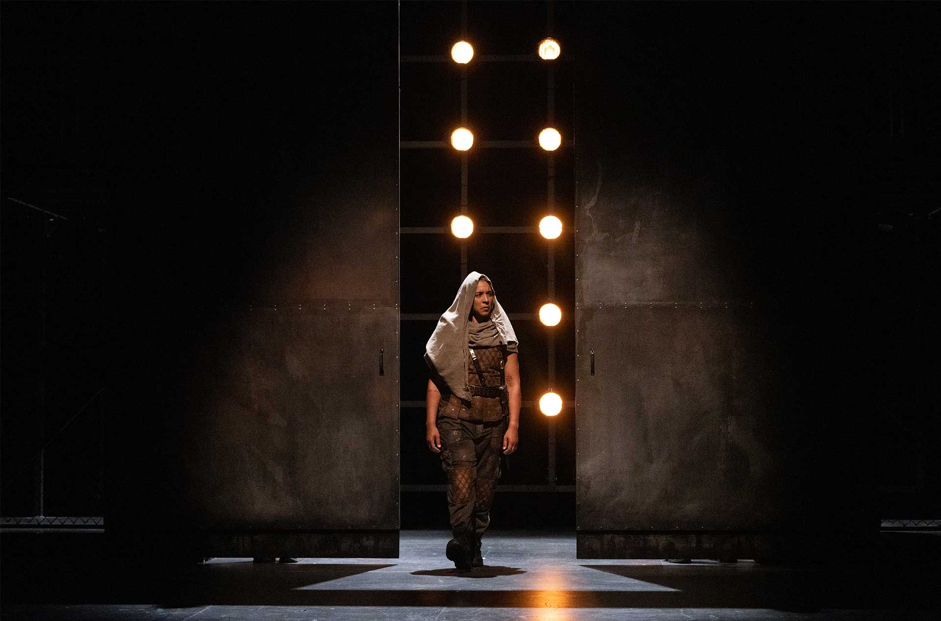 A lone figure dressed in a brown costume with a hooded cloak stands illuminated by a vertical row of bright stage lights. The stark lighting highlights the dramatic entrance through a large dark doorway.