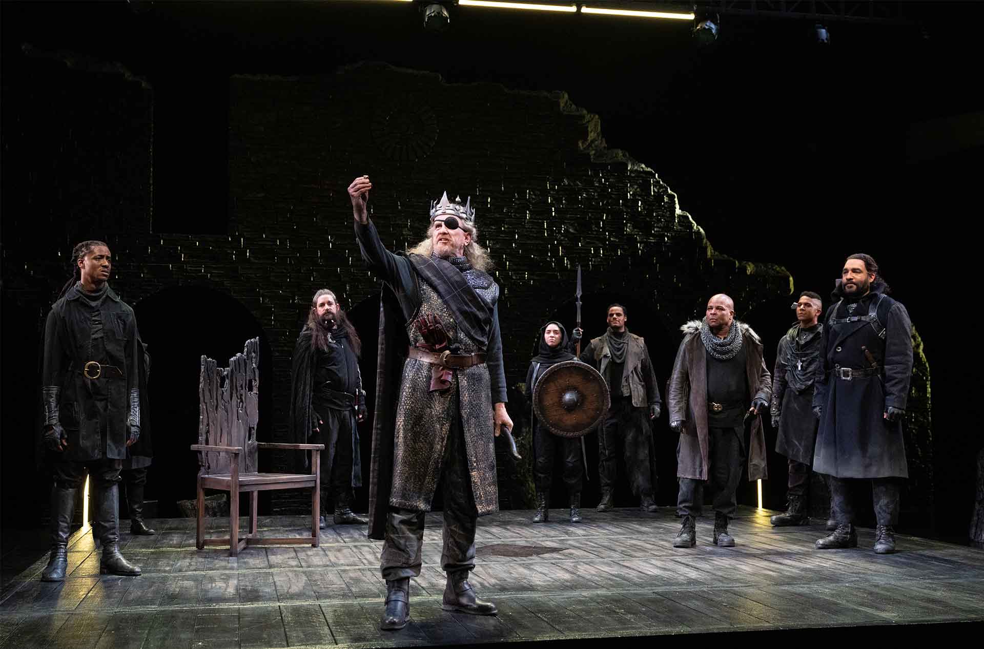 A group of people in medieval-style costumes are on stage. The central figure, wearing a crown and eyepatch, raises one hand as if making a proclamation. The others stand around attentively, with some holding weapons and shields. An ornate wooden chair is positioned to the left of the group.