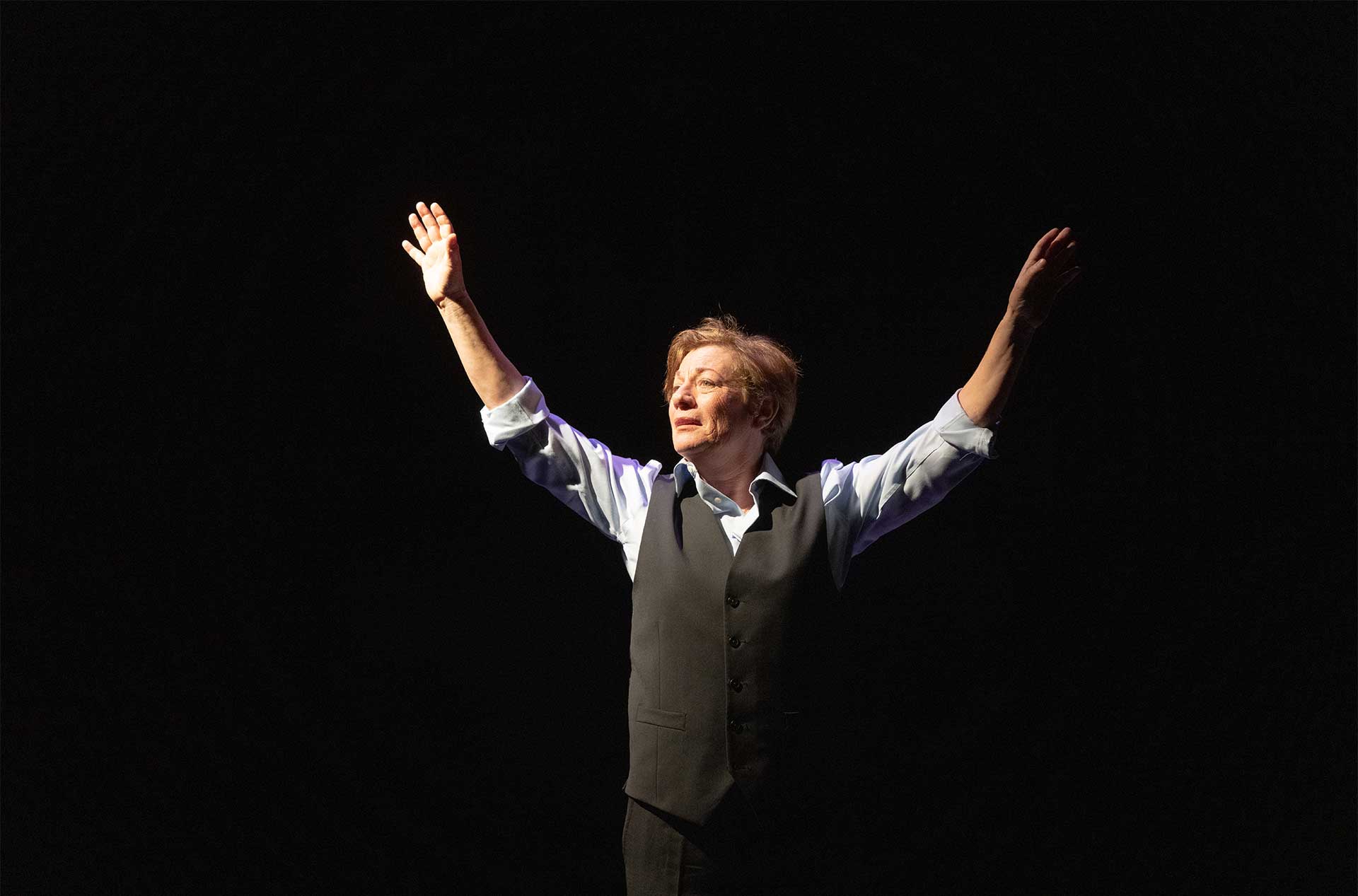 A person standing before a dark background with their hands raised up above their head.
