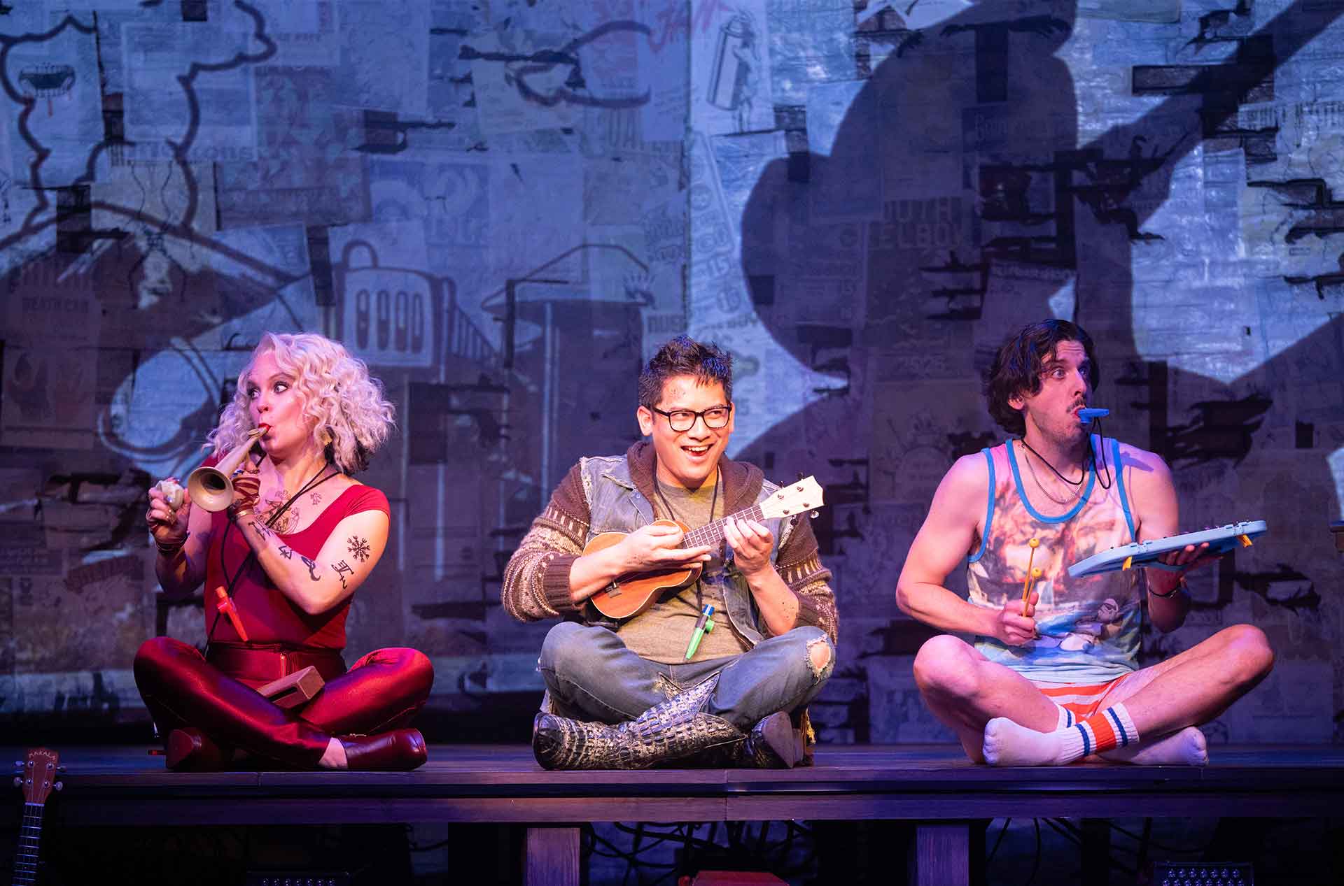 Three performers are seated on stage, each playing a different instrument. The left performer, with blonde hair and a red outfit, plays a trumpet. The middle performer, with glasses and short dark hair, plays a ukulele. The right performer, with curly dark hair and a colorful tank top, plays a toy xylophone and has a kazoo in their mouth. The background features a mural-like urban design.