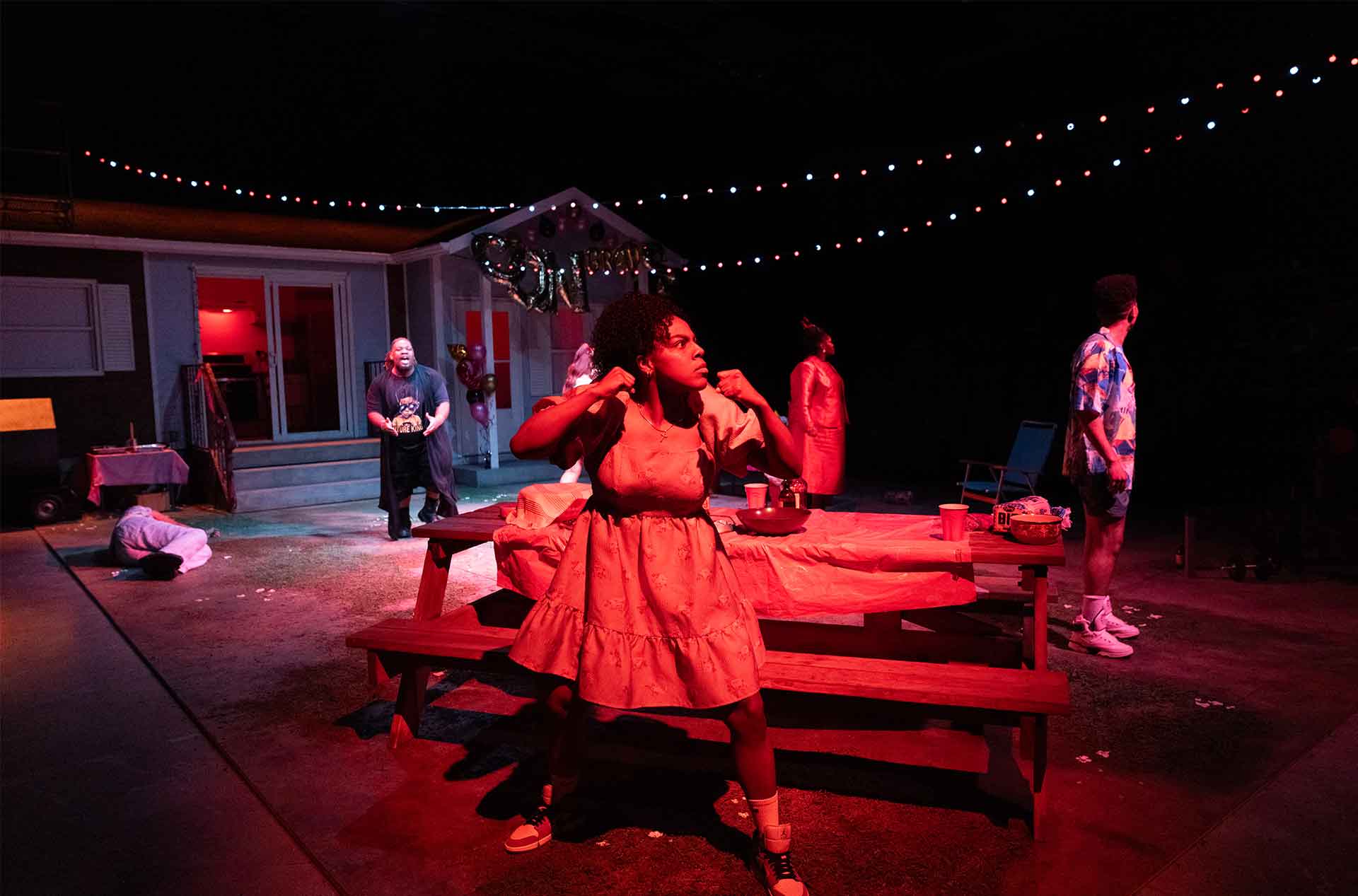 Under dramatic red lighting, a person in a puff-sleeve dress stands in a defensive stance while others react around them in a chaotic backyard scene.