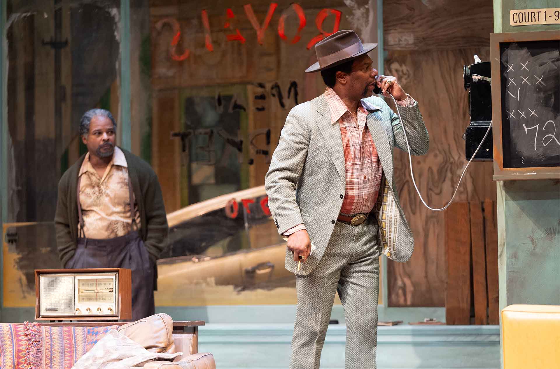 A person speaks urgently into a rotary telephone. Behind them, another person observes the scene with a thoughtful expression.