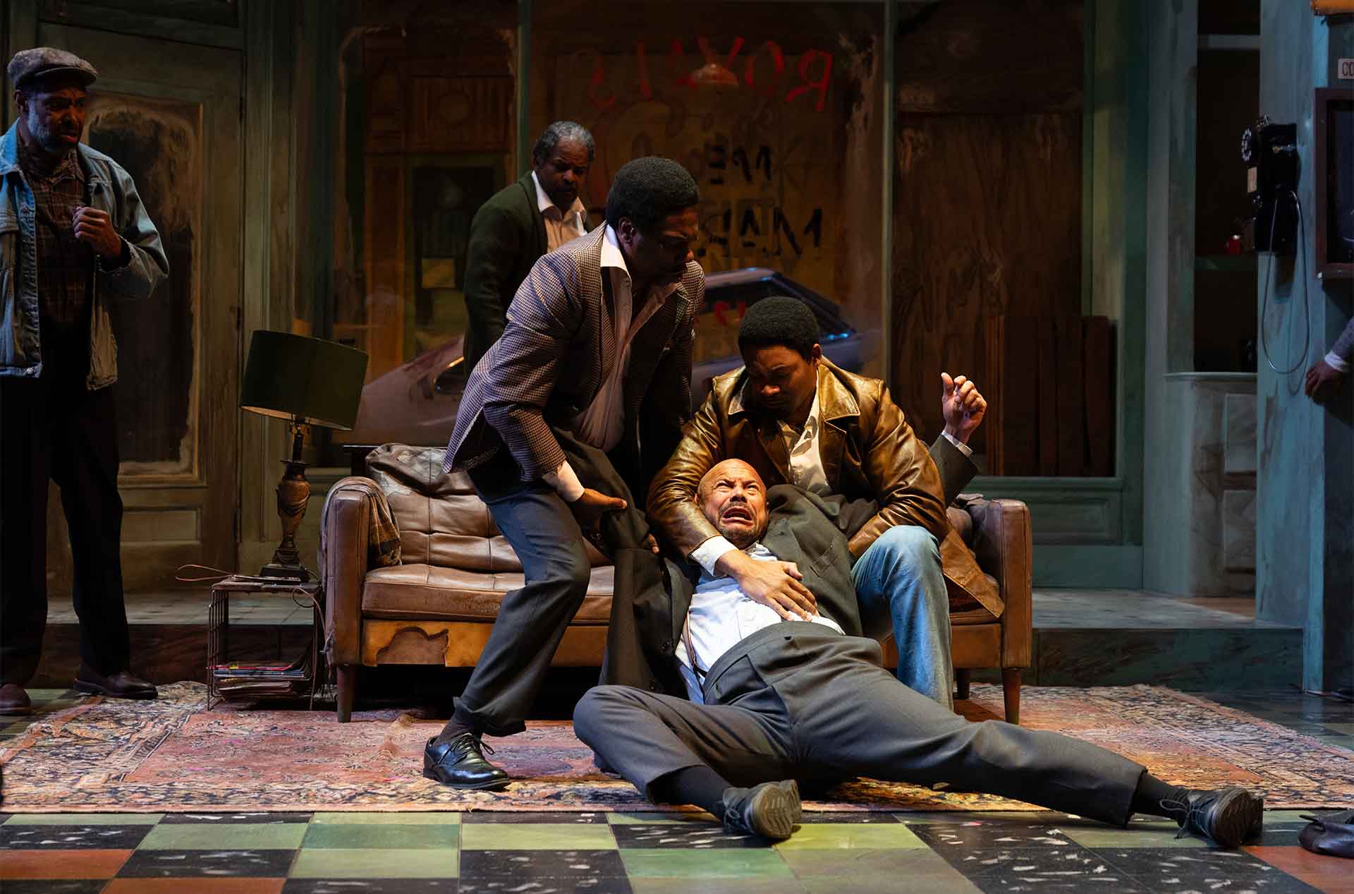 An intense scene unfolds as two people crouch beside a third person who appears to be in distress, lying on the floor with a pained expression