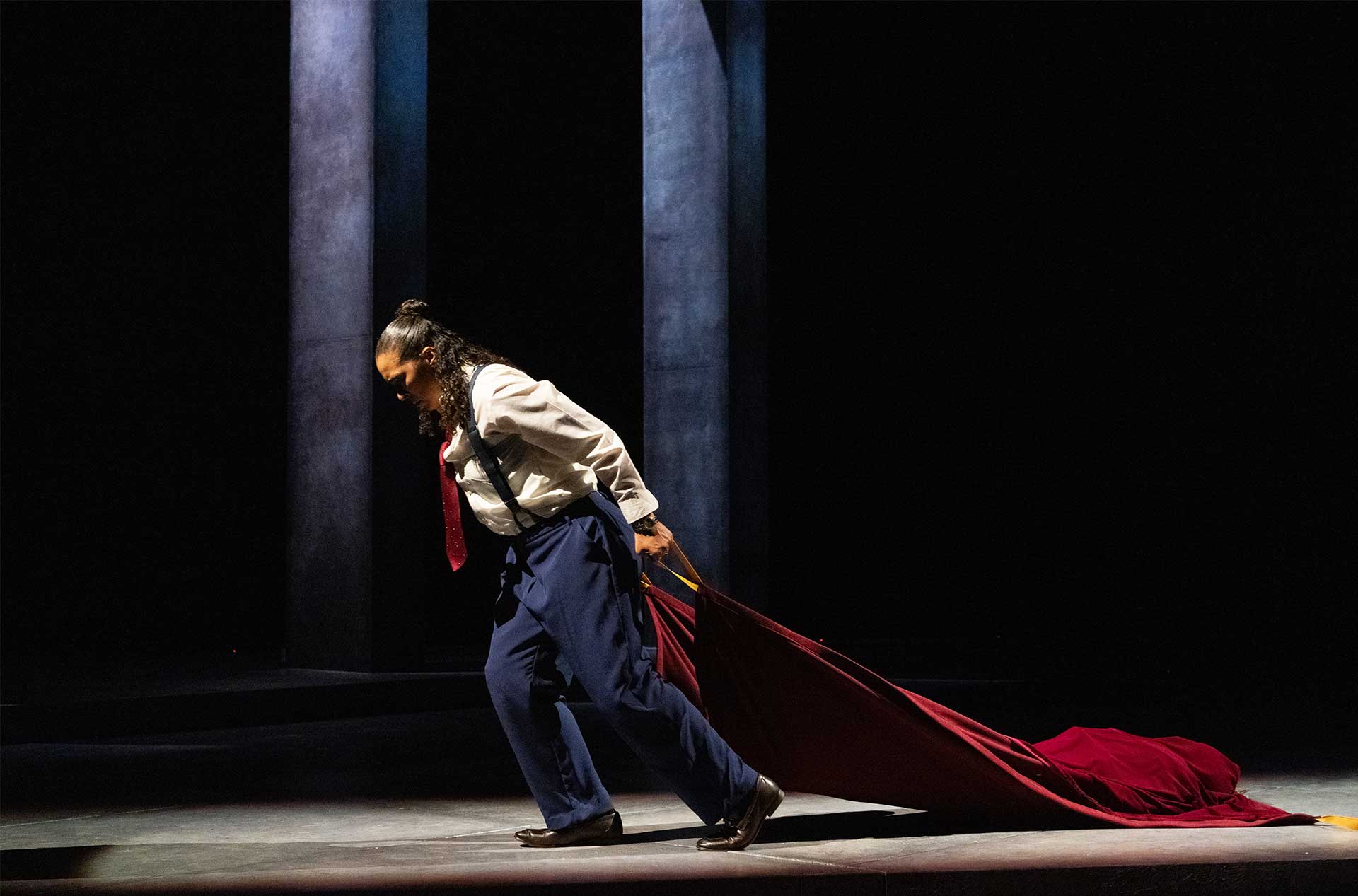 A figure in suspenders and a red tie drags a deep red cloth across a dark stage, their posture strained under the weight.
