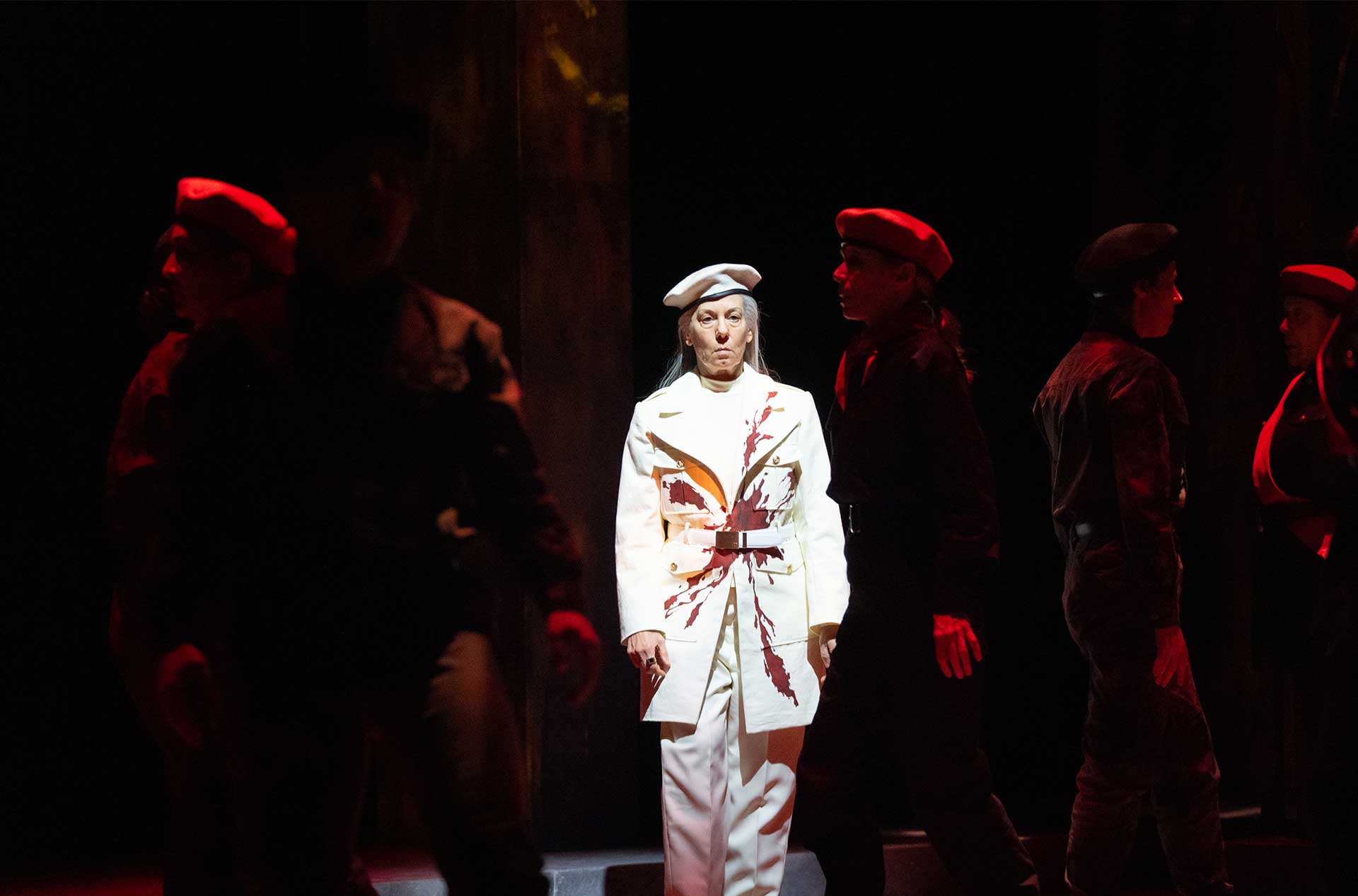 A figure in a white military-style coat with red stains stands under a spotlight, surrounded by others in dark uniforms and red berets in a dimly lit setting.