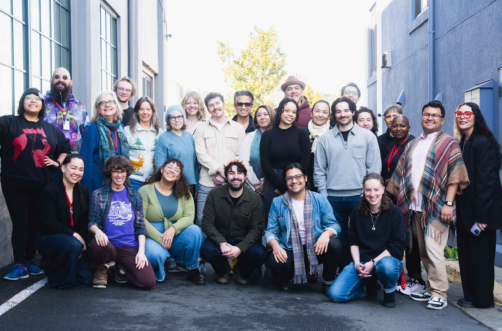 A large group of people poses outside between two buildings, smiling at the camera. They wear a variety of casual and professional clothing, some with lanyards, glasses, or hats.
