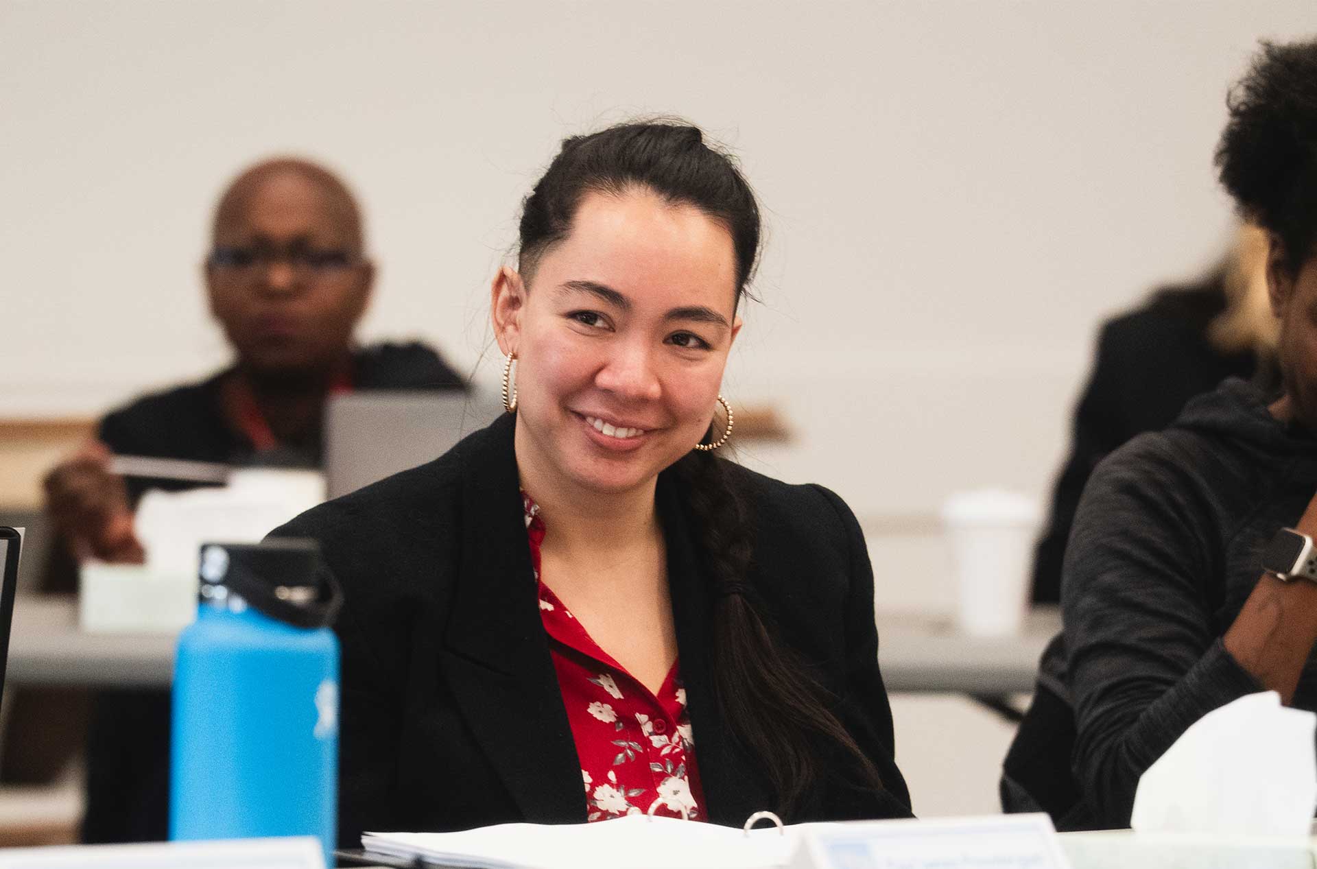A person in a black blazer and red floral shirt sits at a table, smiling attentively. A blue water bottle is in front of them, and other attendees are visible in the background.