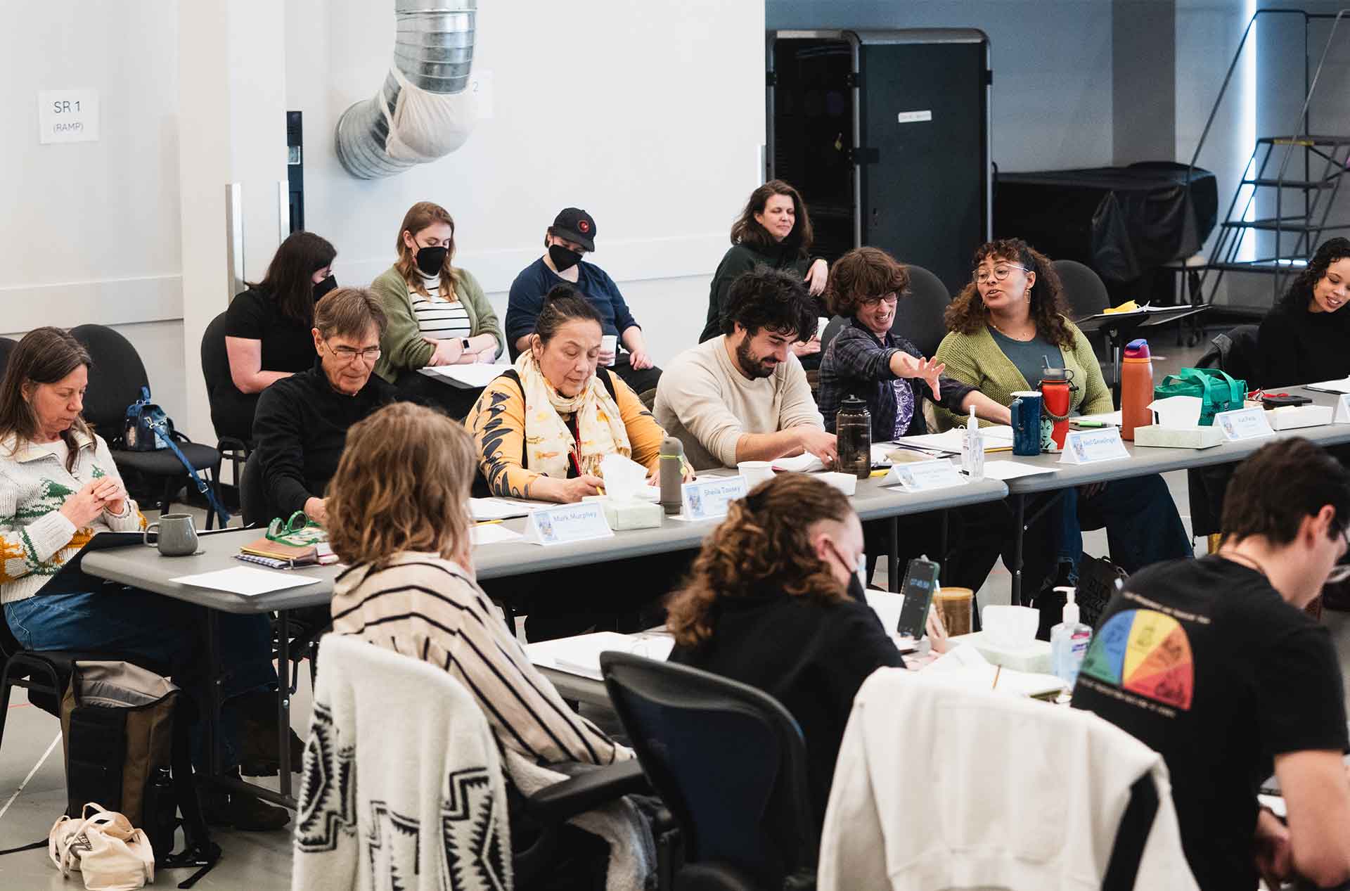 A group of people sits around a long table, engaged in discussion or taking notes. Some are speaking animatedly, while others are focused on their papers or devices.