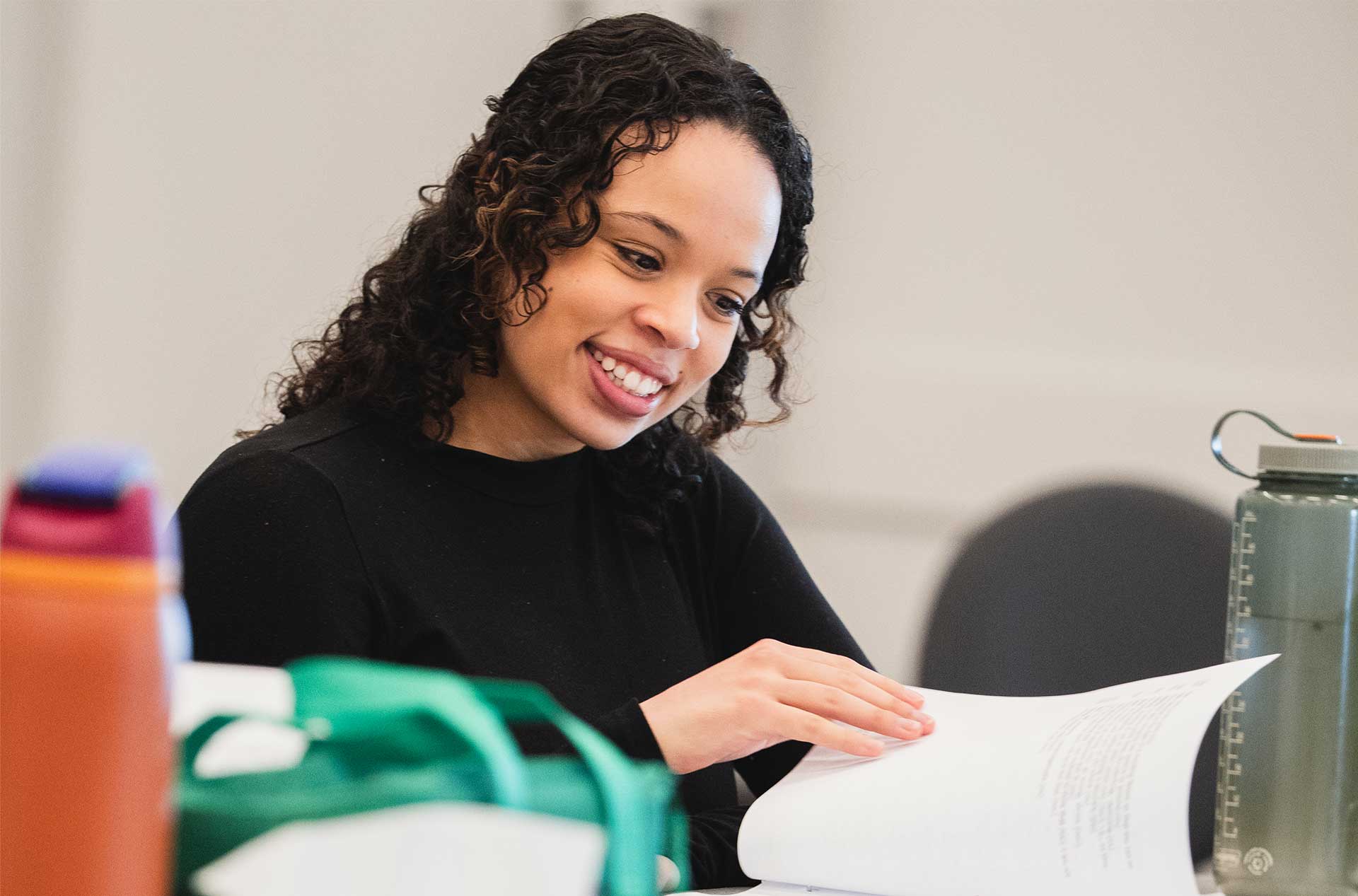 A person in a black shirt smiles while flipping through pages of a script. A green water bottle and bags are on the table beside them.