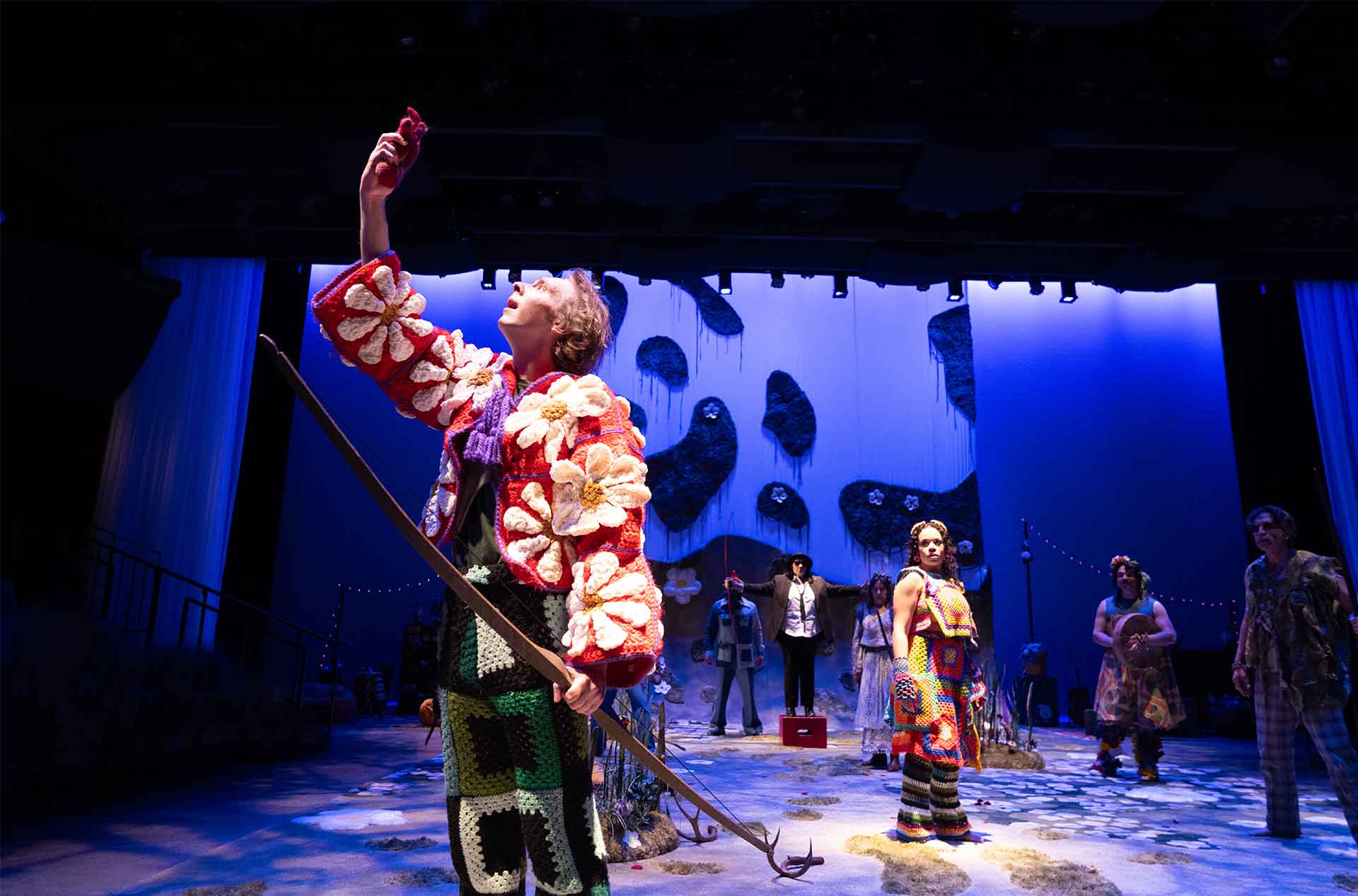 A barefoot performer in a flower-covered outfit raises an object dramatically, as others look on under moody lighting with a surreal, mossy backdrop.