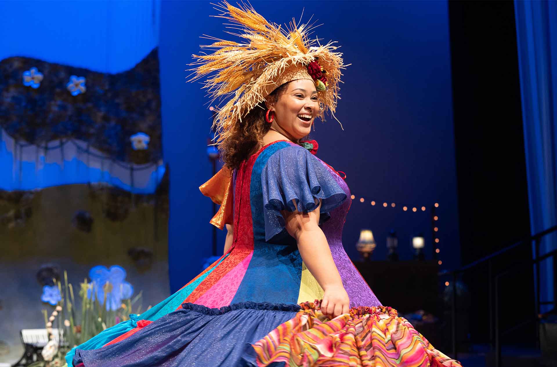 A smiling performer twirls in a sparkling, patchwork dress with puffed sleeves and a straw headdress, surrounded by whimsical stage lighting and paper flowers.