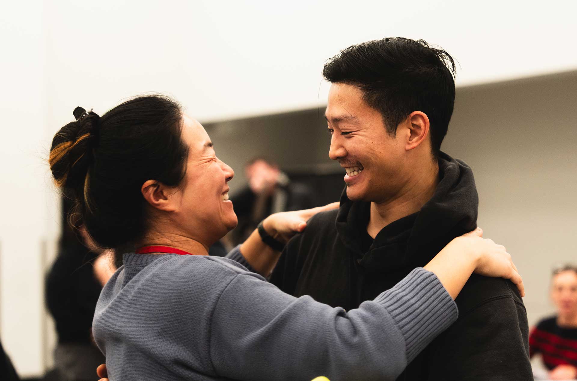 Two individuals share a warm moment, smiling and holding hands on each other’s shoulders during a break in rehearsal. The background is blurred, emphasizing their connection.