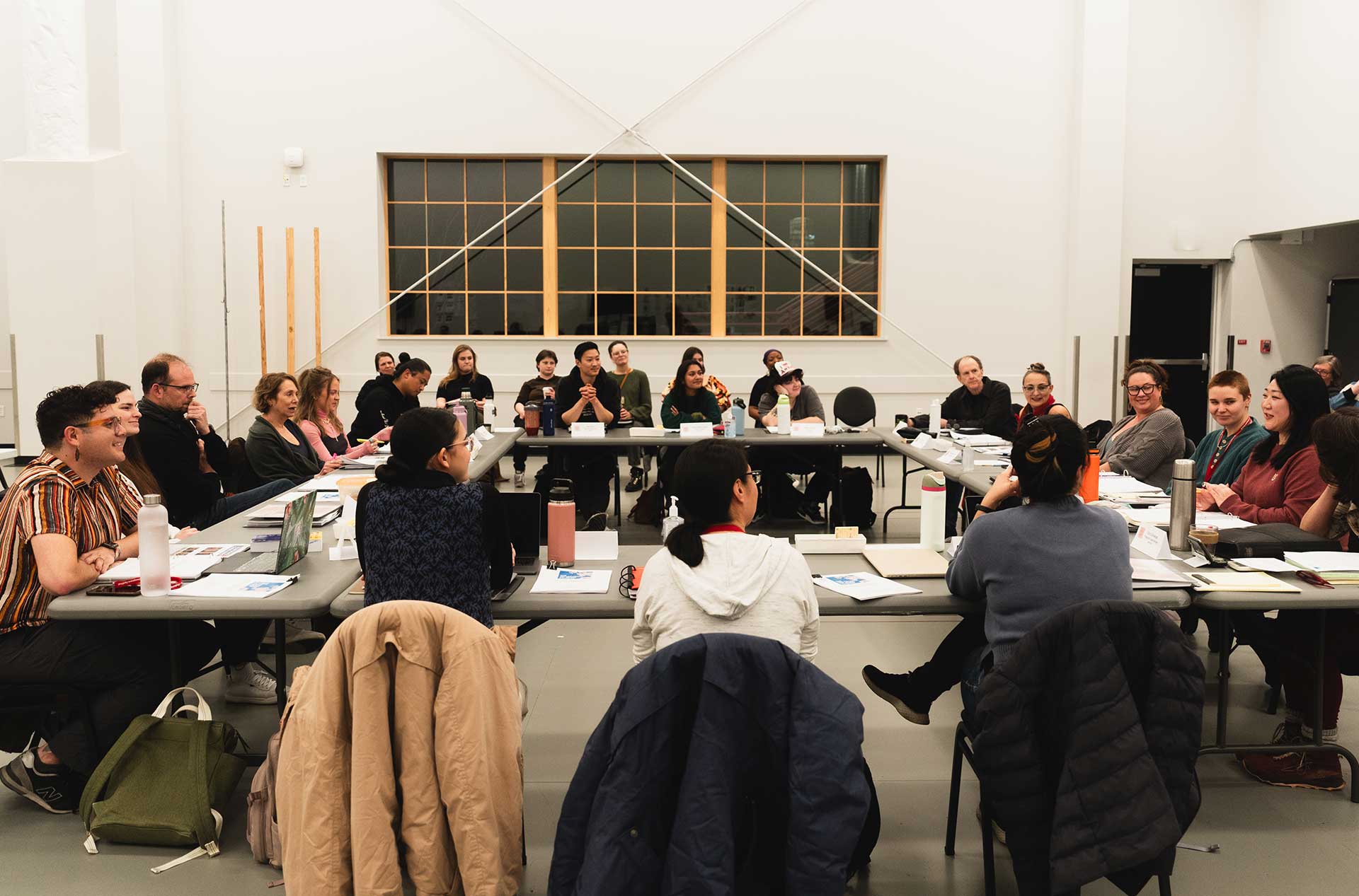 A wide shot of a rehearsal room, with participants seated in a square table arrangement. The room is brightly lit, with a large window at the far end and participants actively engaged in discussion.