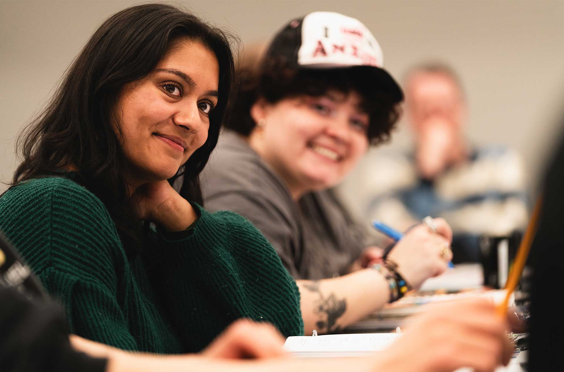 A person wearing a green sweater glances toward the camera with a slight smile during a rehearsal. Others around the table appear focused, with one person in a cap smiling broadly in the background.