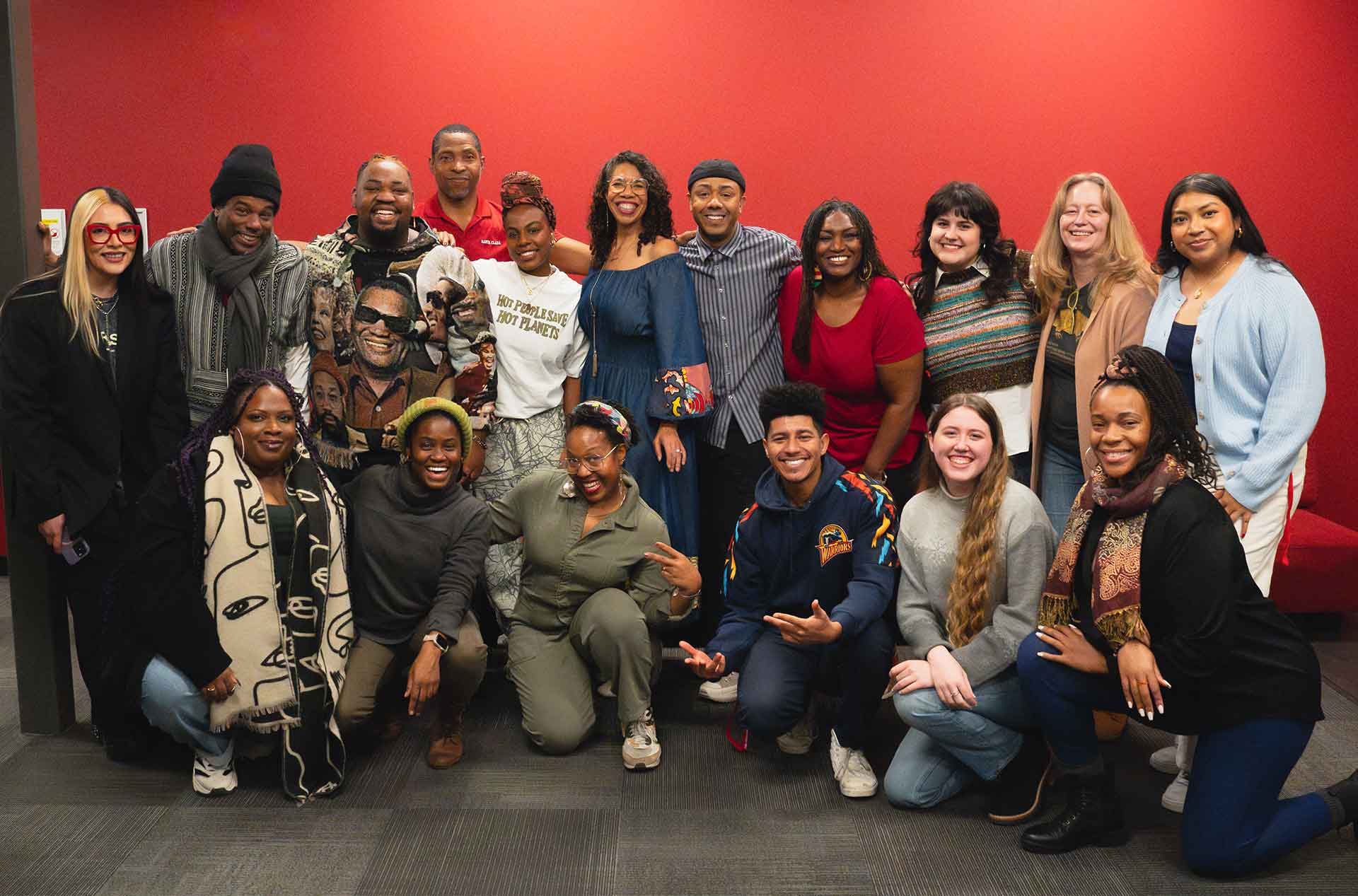 The cast and creative team posing together in front of a red wall. They are smiling, with some crouching in front and others standing behind, wearing a mix of casual and expressive clothing.