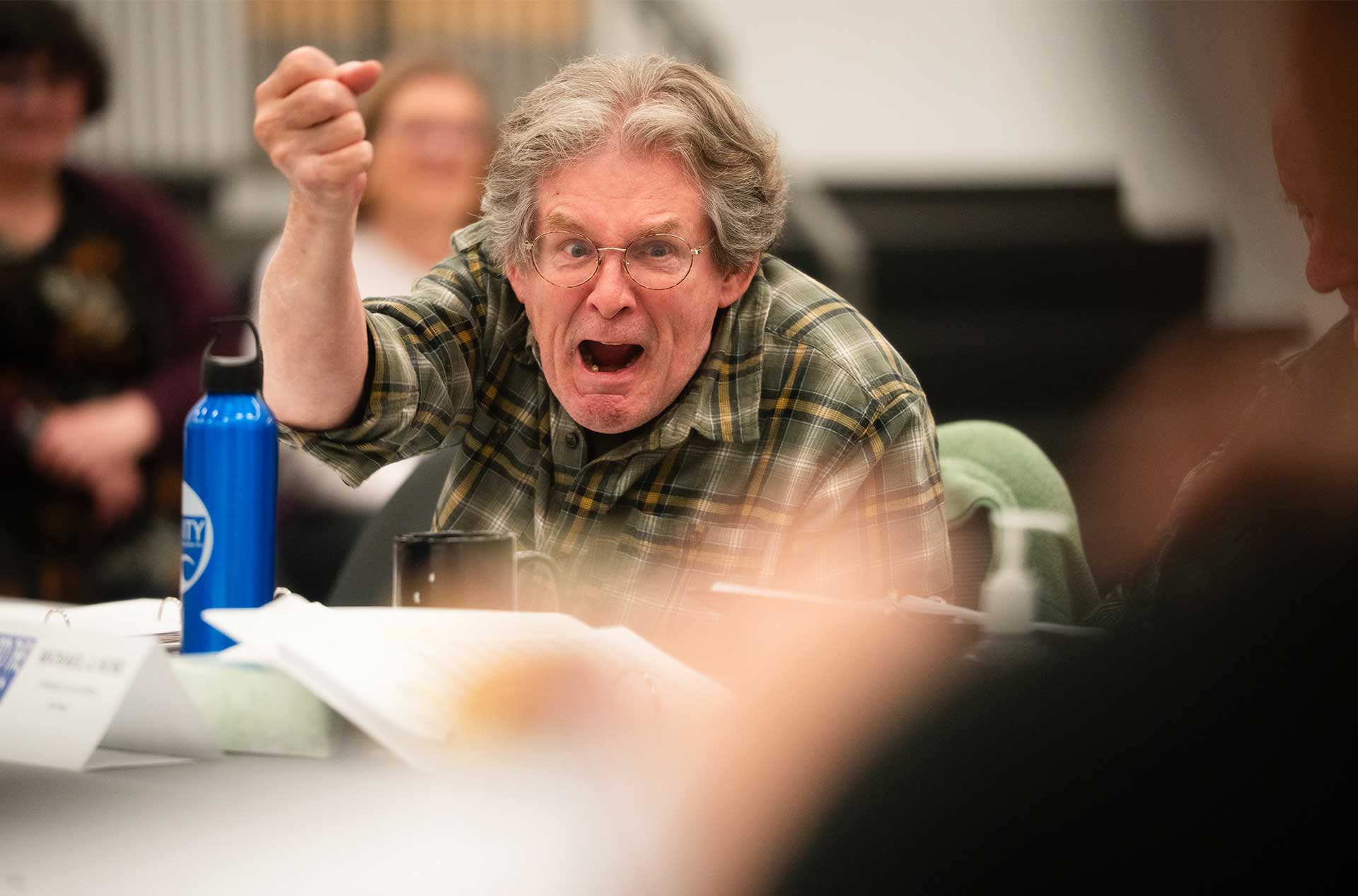 A person in a plaid shirt energetically points and shouts during an expressive moment in rehearsal, surrounded by scripts and water bottles.