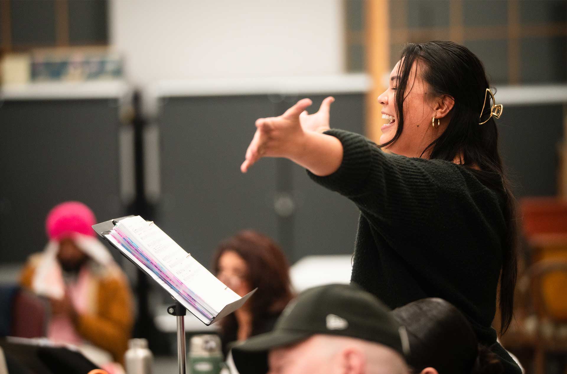 A person extends their arm dramatically while standing behind a music stand filled with color-coded sheets during a performance rehearsal.