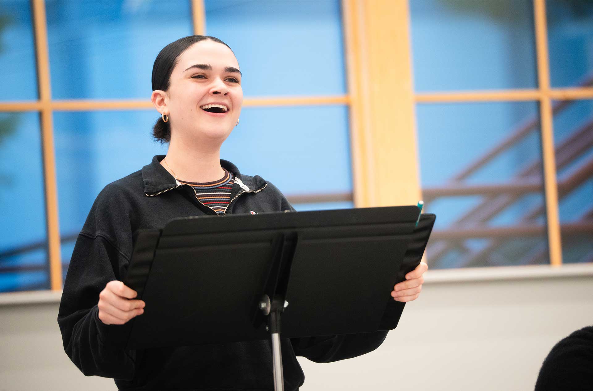 A smiling person stands behind a music stand while rehearsing lines, with a large window in the background.