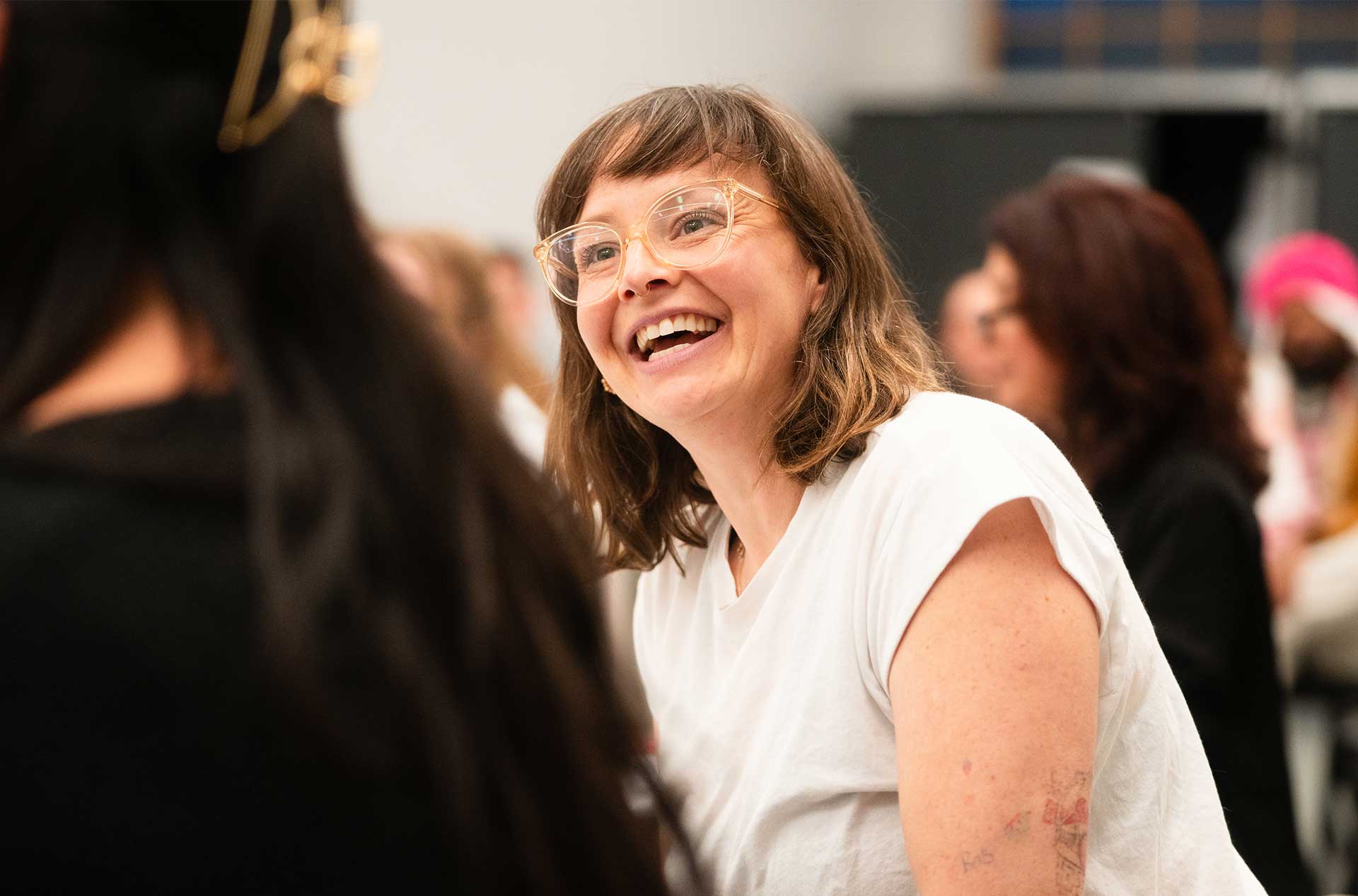 A person with shoulder-length hair and glasses smiles warmly while seated among others during rehearsal.