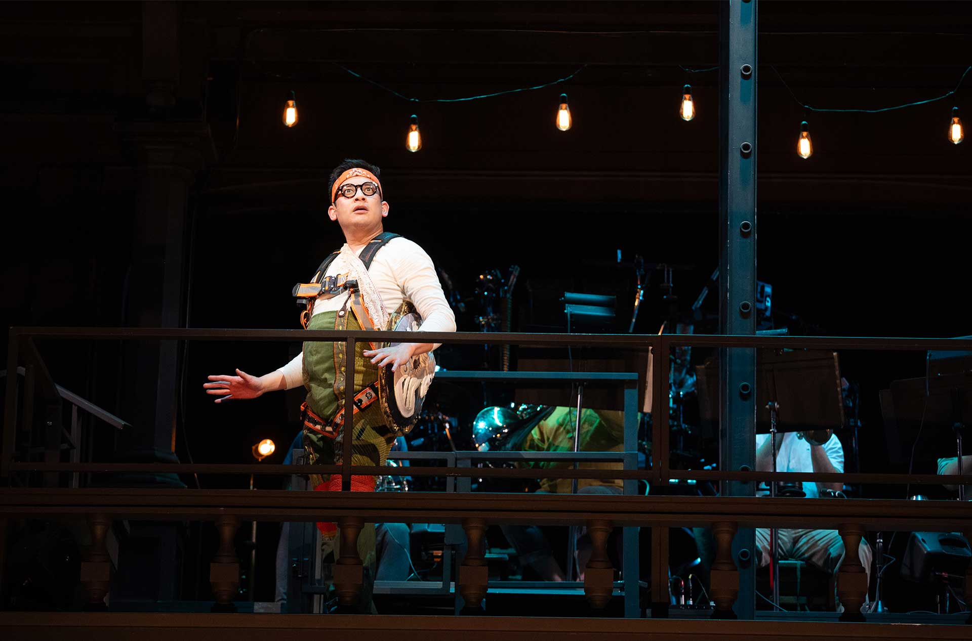 An actor stands dramatically on an upper platform, lit by string bulbs, wearing glasses and a costume with climbing gear elements, looking upwards in suspense or wonder.