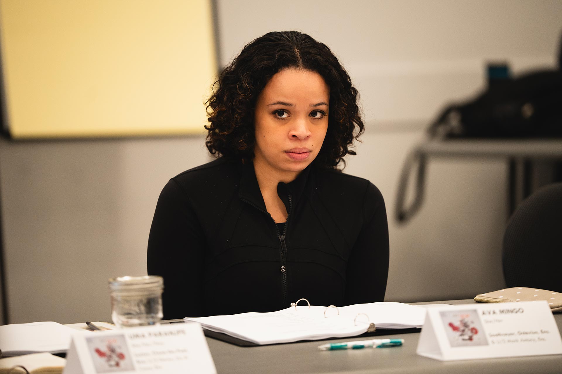 A cast member sits with a focused expression during the rehearsal. A binder and pen are in front of them, with a nameplate indicating their role.