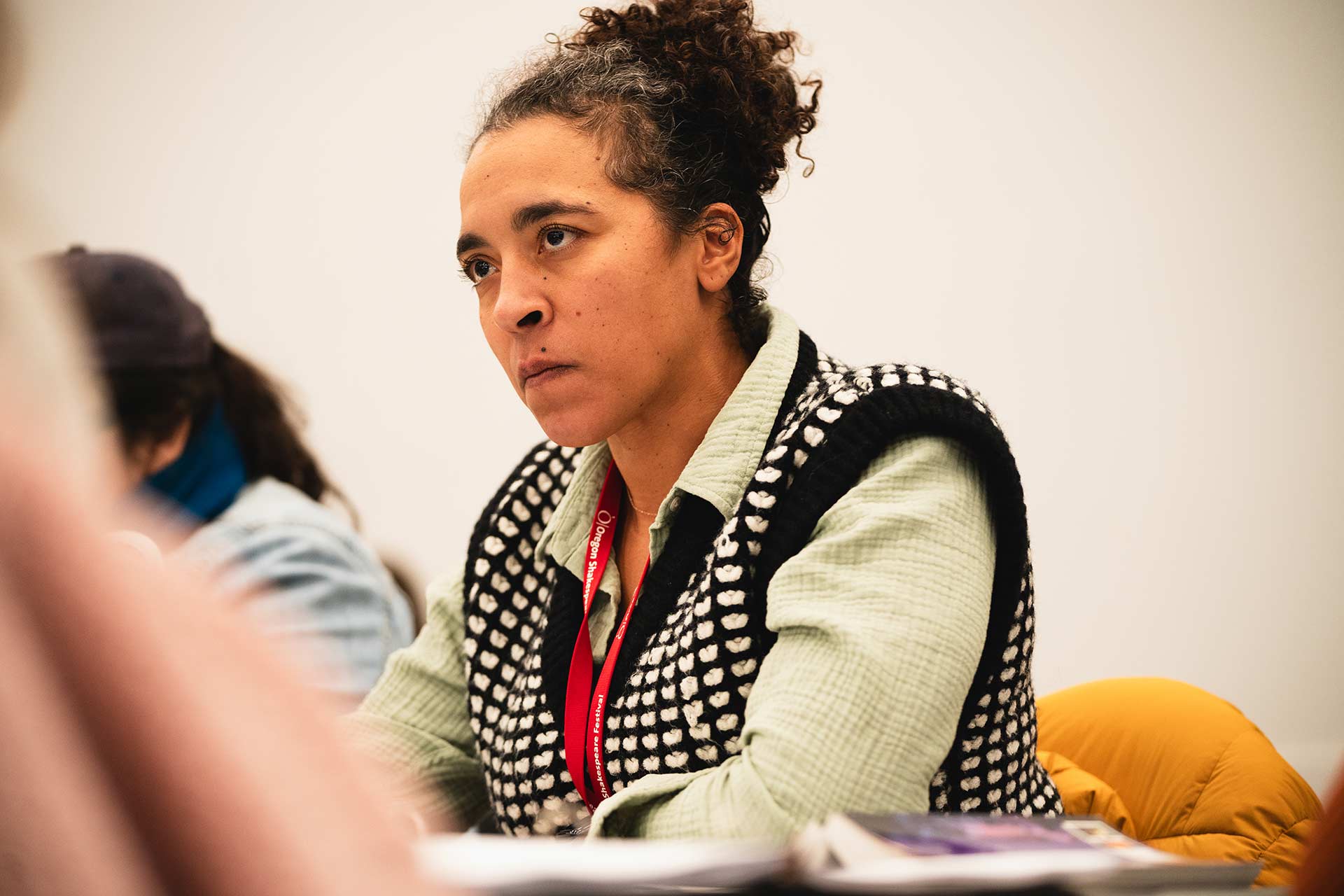 A rehearsal participant sits attentively, focusing on the discussion. They are wearing a patterned vest and a lanyard with a badge, with scripts and notes visible in front of them on the table.