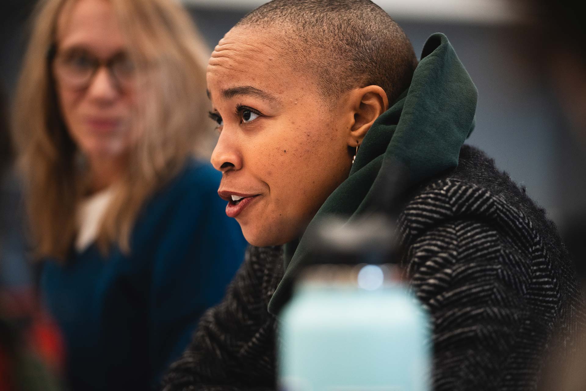 A close-up of a cast member speaking during a rehearsal discussion. The individual sits at a table, with another person visible in the background.