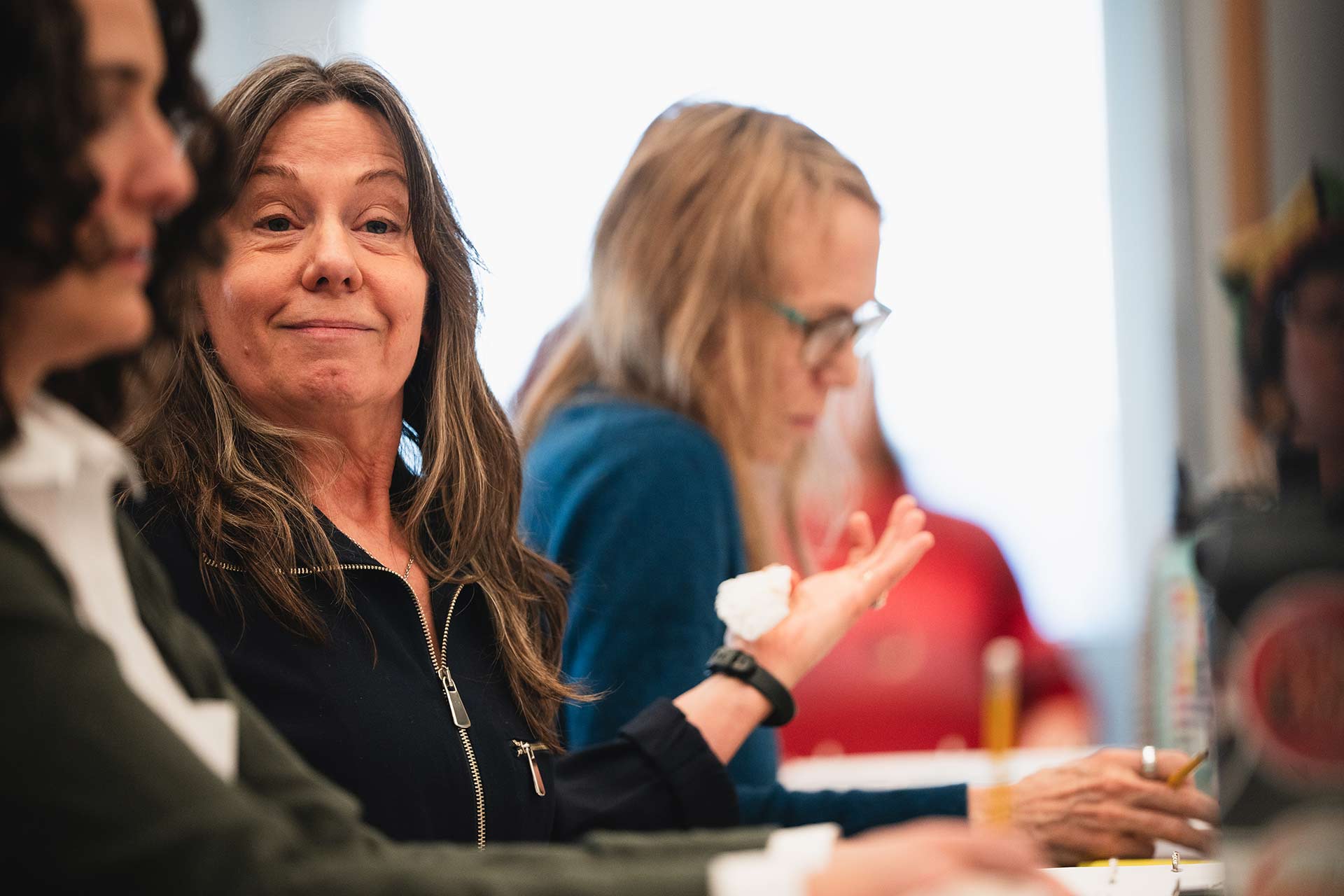 A rehearsal participant gestures with one hand while speaking, smiling as they engage in discussion. Two others sit beside them, reviewing their scripts and taking notes.
