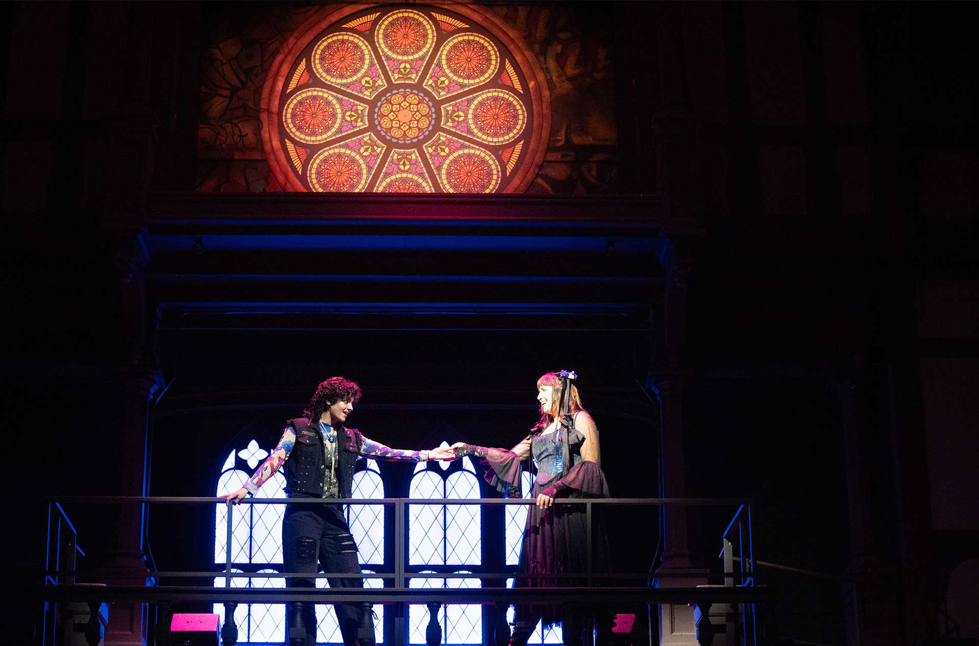 Two performers in modern punk-inspired costumes stand on a balcony, reaching toward each other under a glowing circular stained glass window, evoking a romantic or dramatic moment.
