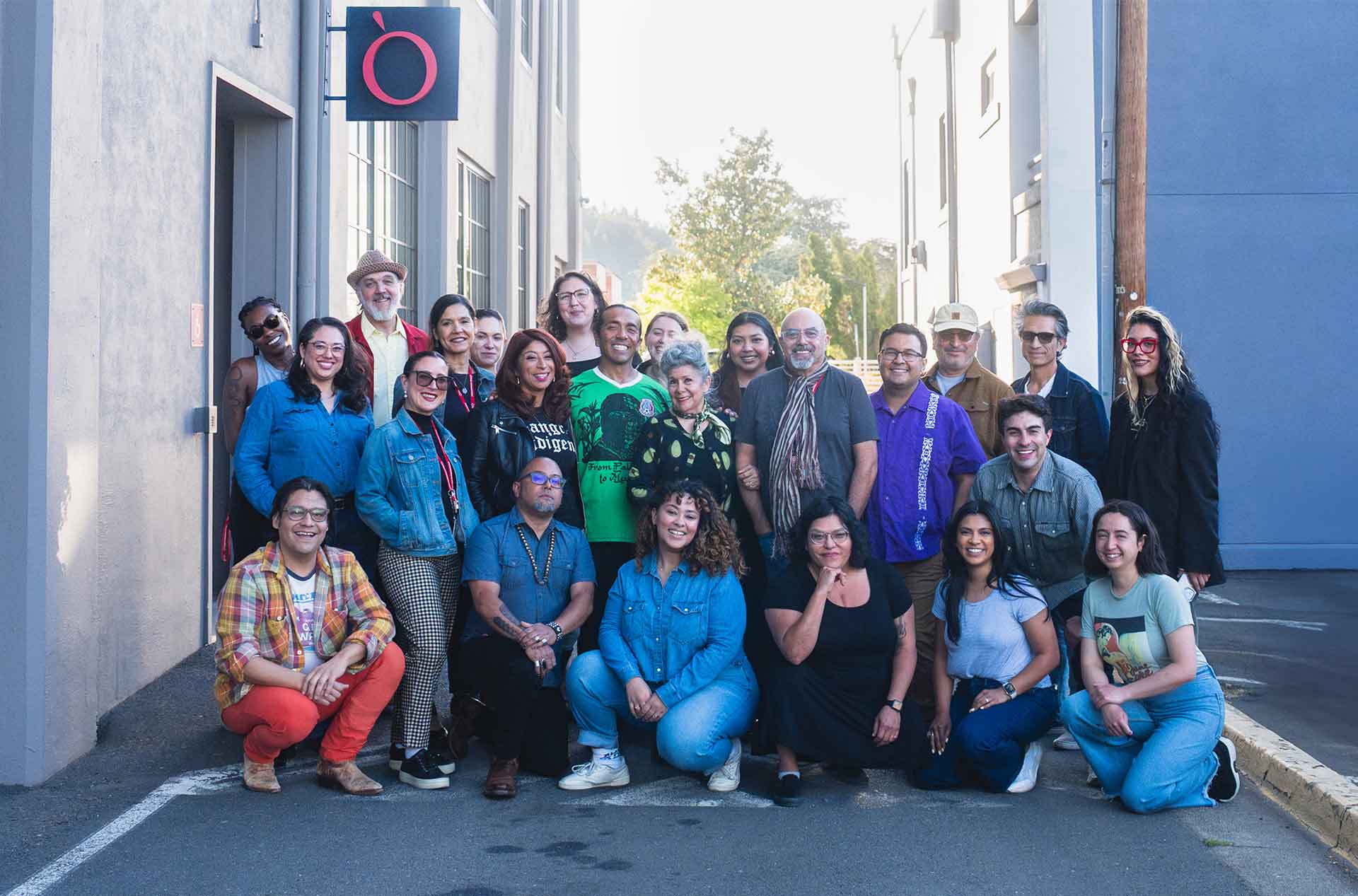 A group photo of cast and crew standing and kneeling outside a rehearsal space, smiling beneath a sign with a red O.