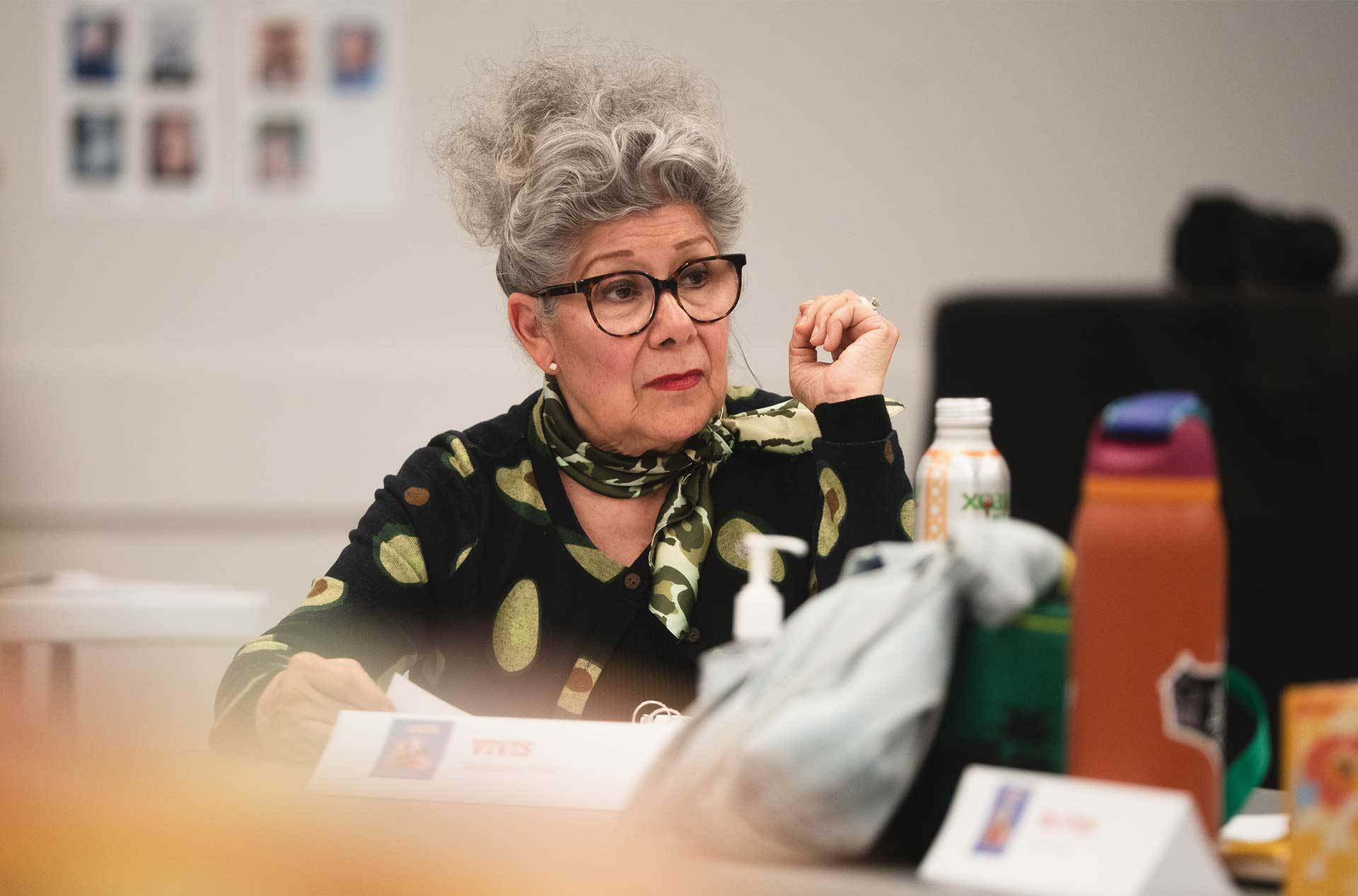 A rehearsal participant sits at a table listening attentively, surrounded by colorful water bottles, a scarf, and a visible nameplate.