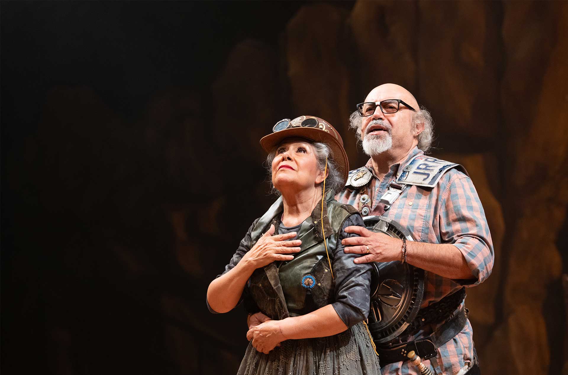 Two actors stand closely, one with hand over heart and the other embracing from behind, both wearing costume pieces resembling armor.