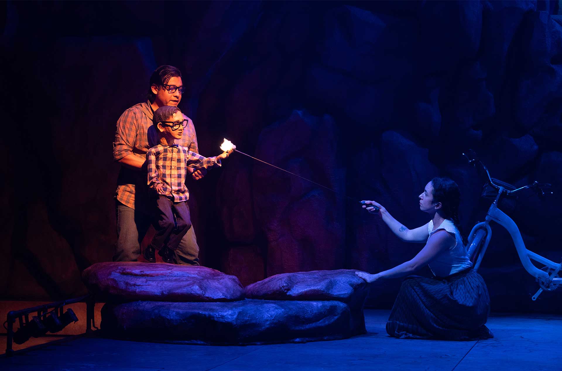 Puppeteer holding a child puppet roasting a marshmallow over a stick, while another performer kneels across from them in blue stage lighting.