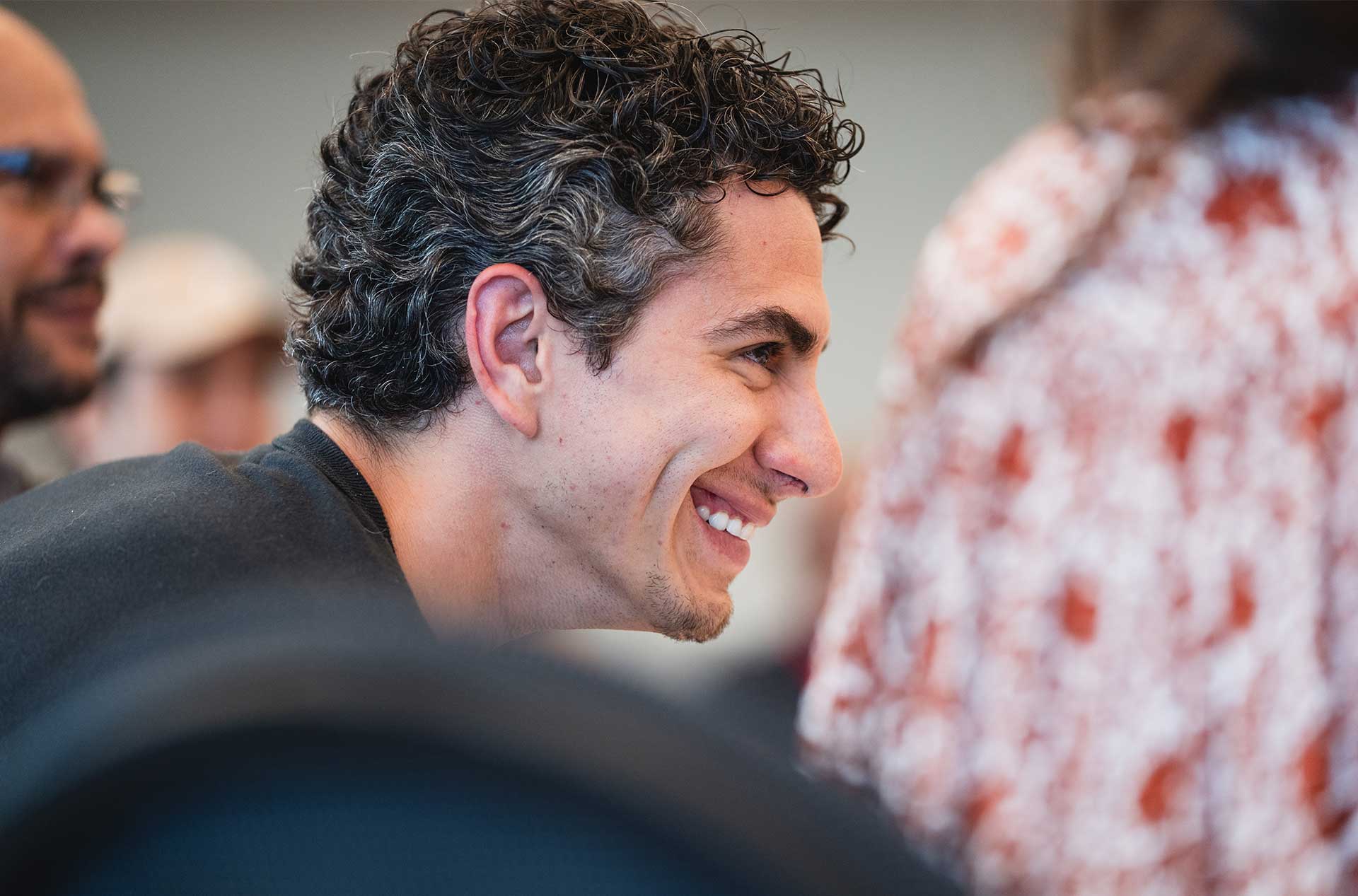 A close-up profile of a smiling person with short curly hair and light stubble, surrounded by blurred faces and patterned clothing in the foreground.