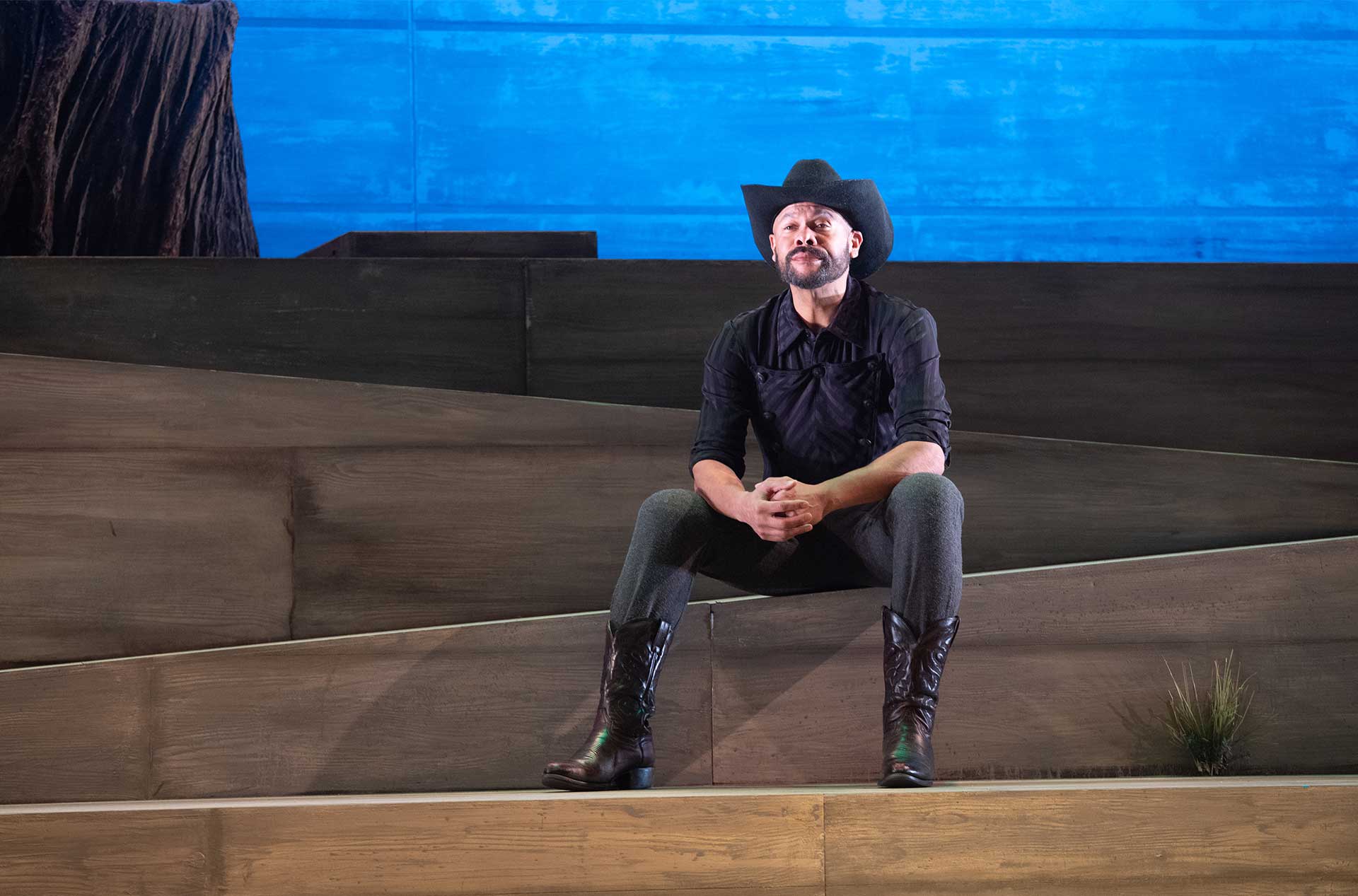 A performer in cowboy boots and a black shirt sits on a stepped wooden set, lit against a bright blue background.